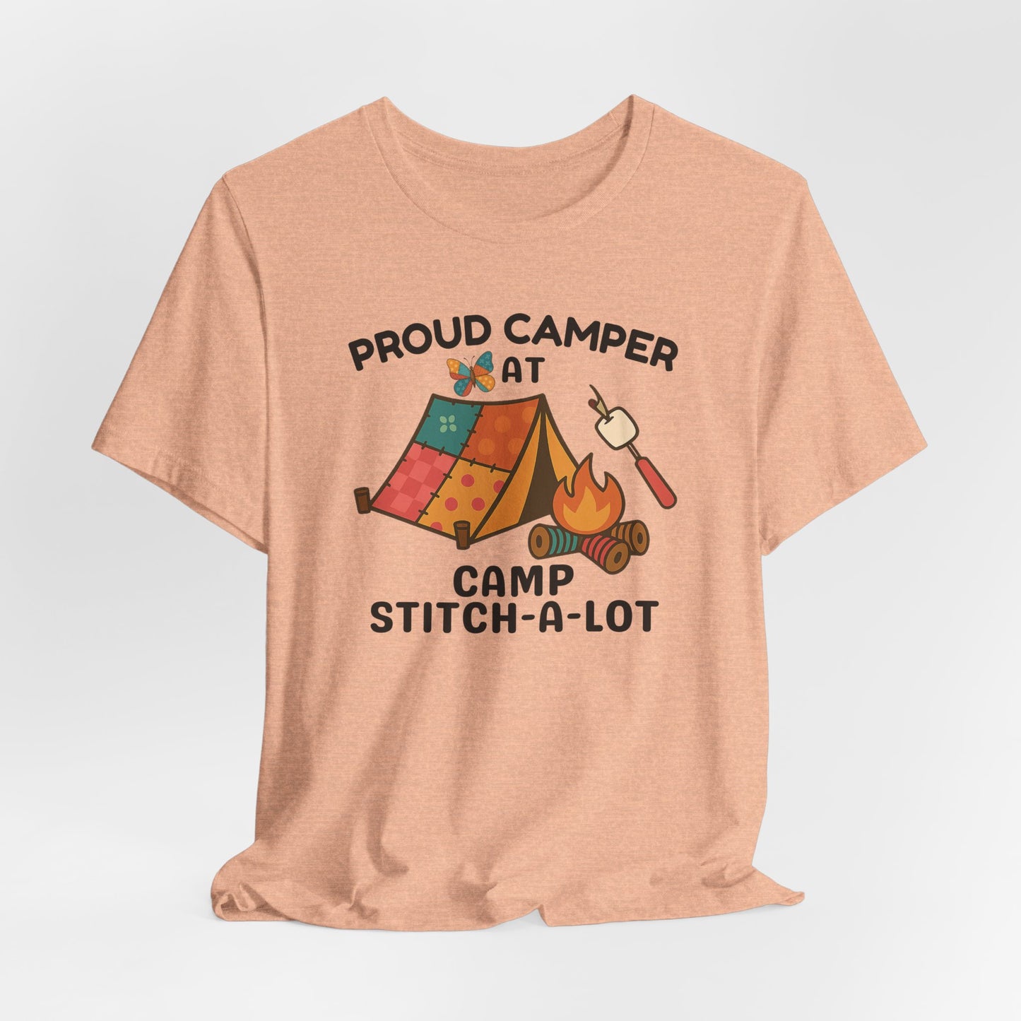 A A Heather Peach t-shirt featuring a colorful design with a patchwork tent, thread spool campfire, marshmallow on a seam ripper stick, and a butterfly. Text reads "Proud Camper at Camp Stitch-a-Lot." Fun gift for quilting retreats.