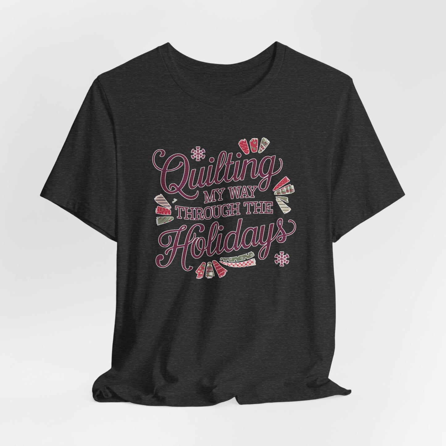 A Dark Grey Heather playful holiday-themed t-shirt with the phrase "Quilting My Way Through the Holidays" surrounded by quilting-inspired decorations like fabric swatches and snowflakes.
