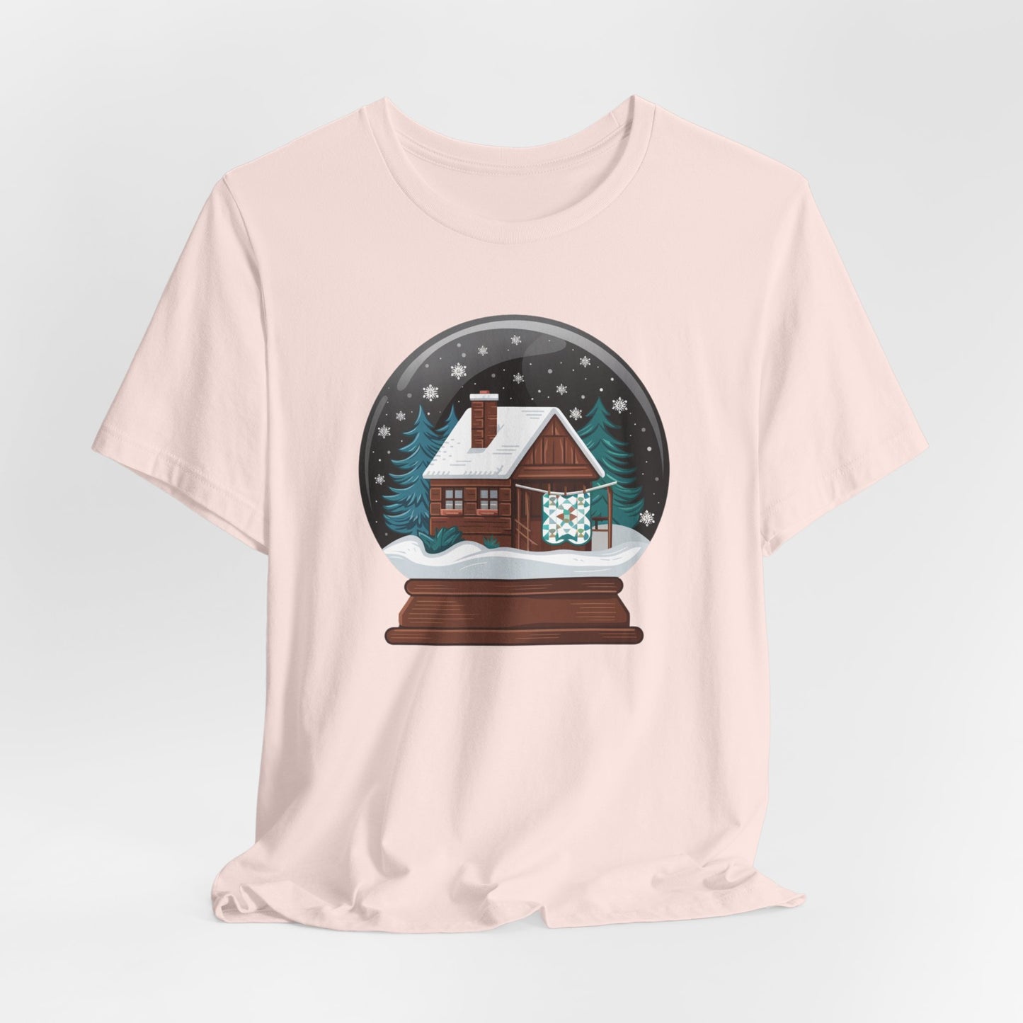 A Soft Pink christmas quilting t-shirt featuring a cozy log cabin covered in snow, with a quilt hanging outside, set inside a snow globe with swirling snowflakes, blending quilting and holiday warmth.