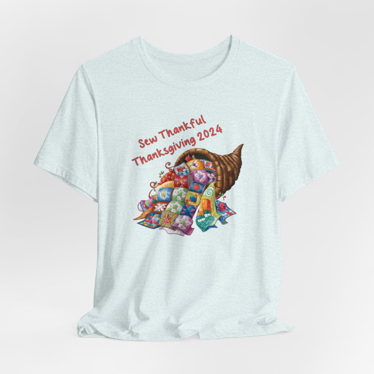 A Heather Ice Blue thanksgiving quilting t-shirt featuring a colorful quilted cornucopia overflowing with various quilt patterns and designs. Text reads "Sew Thankful Thanksgiving 2024" in red. Vibrant illustration combines quilting motifs with Thanksgiving imagery.