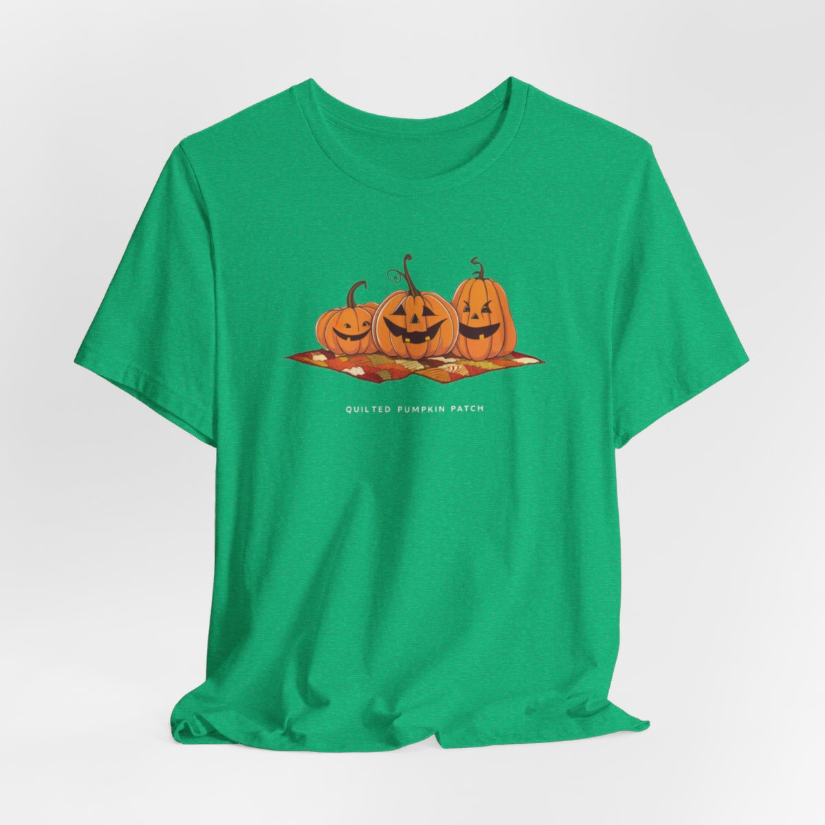 A Heather Kelly T-shirt with three smiling jack-o'-lanterns sitting on a colorful autumn-themed quilt. The pumpkins have carved faces with various expressions, and the quilt beneath them features a patchwork of fall colors. Text below reads "QUILTED PUMPKIN PATCH".