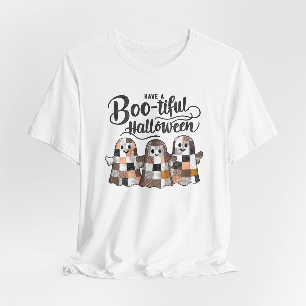 A White Halloween Quilting T-Shirt featuring three smiling patchwork ghosts made of various quilt patterns in autumn colors. Text reads "Have a Boo-tiful Halloween" in stylized lettering. Perfect for quilters who love Halloween.