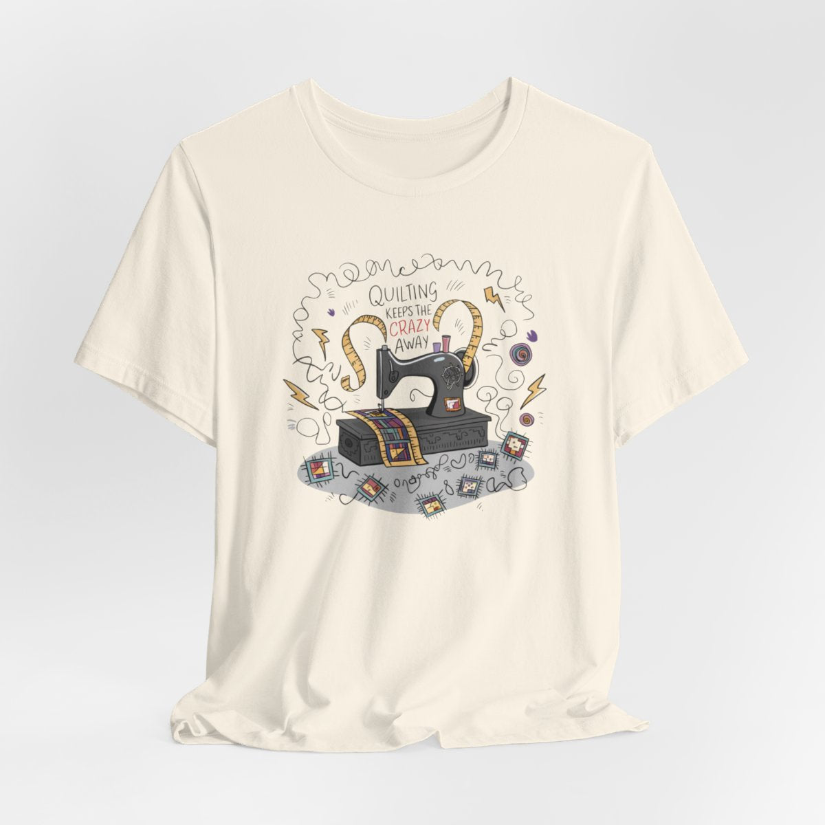 A Natural T-Shirt with a vintage sewing machine, measuring tape heart, and text "Quilting Keeps the Crazy Away" amidst swirls and quilting motifs.