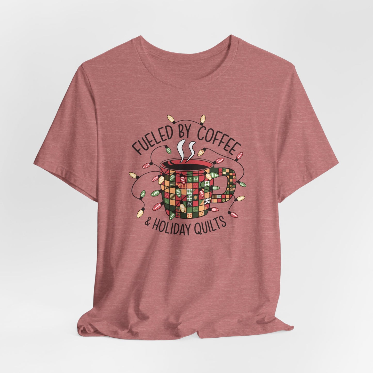 A Heather Mauve christmas quilting t-shirt featuring a cozy coffee mug covered in patchwork quilt patterns, wrapped in festive holiday lights, with the phrase "Fueled by Coffee & Holiday Quilts" surrounding the design.