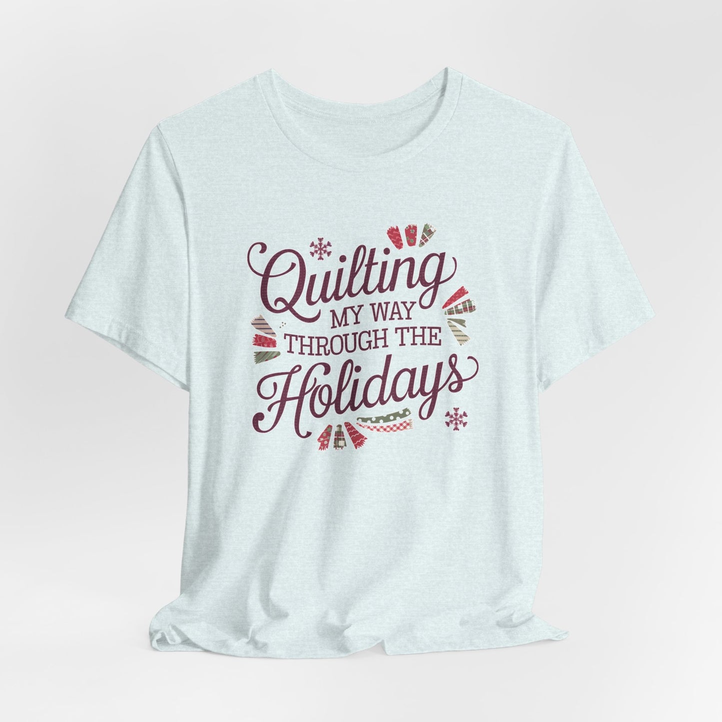 A Heather Ice Blue playful holiday-themed t-shirt with the phrase "Quilting My Way Through the Holidays" surrounded by quilting-inspired decorations like fabric swatches and snowflakes.