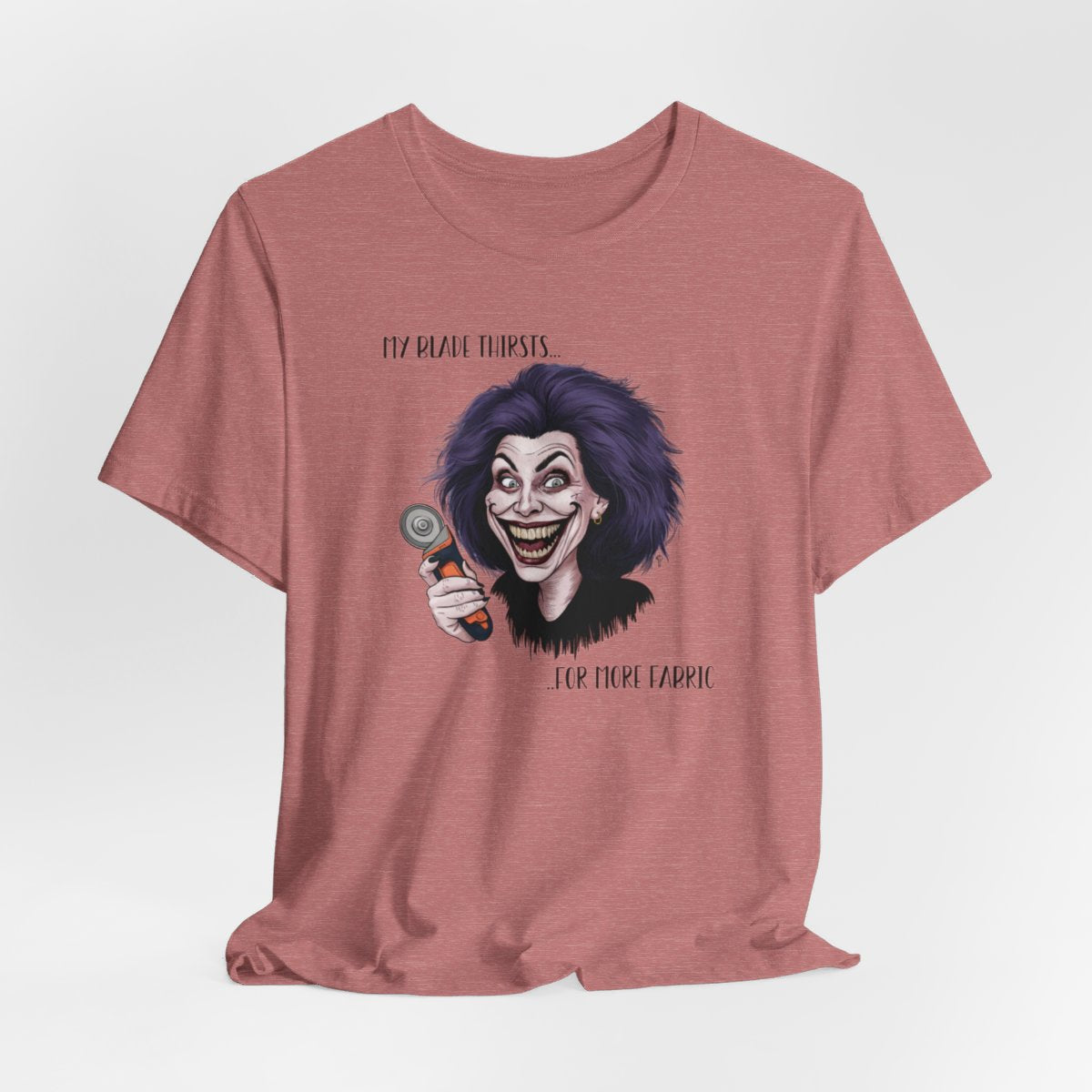 A Heather Mauve T-shirt showing a cartoonish character with wild purple hair and an exaggerated grin, holding a rotary cutter. Text reads "My blade thirsts... for more fabric", humorously depicting a quilter's endless desire for new materials.