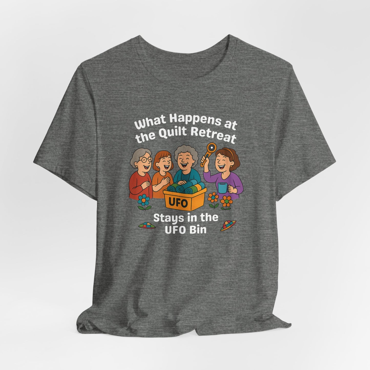 A A Deep Heather t-shirt featuring a cartoon design of four laughing women at a quilt retreat with a bin labeled “UFO” full of fabric. Surrounded by patchwork flying saucers and flowers. Text reads “What happens at the quilt retreat stays in the UFO bin.”