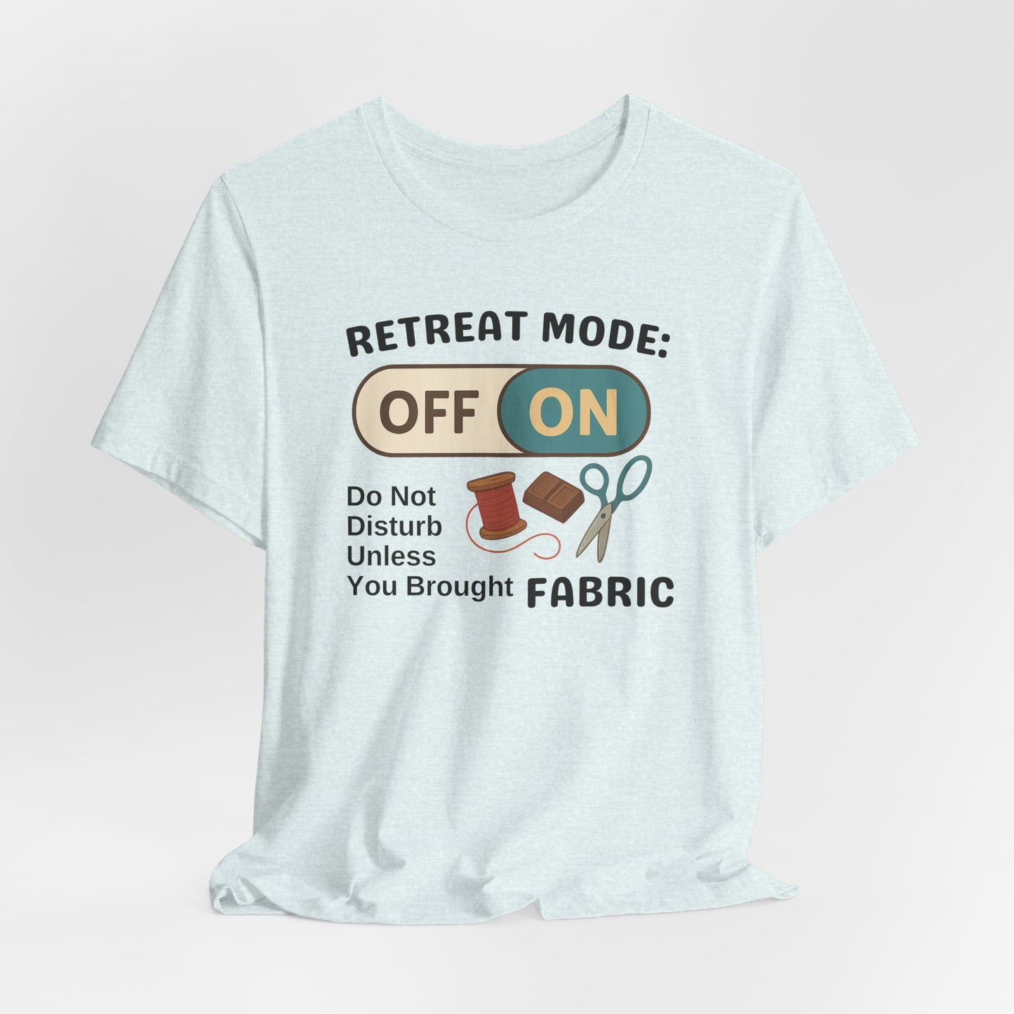 A A Heather Ice Blue t-shirt with a toggle switch that says "Retreat Mode: OFF/ON" surrounded by a spool of thread, chocolate, and scissors. Text below reads "Do not disturb unless you brought fabric." Fun gift for quilting retreats.