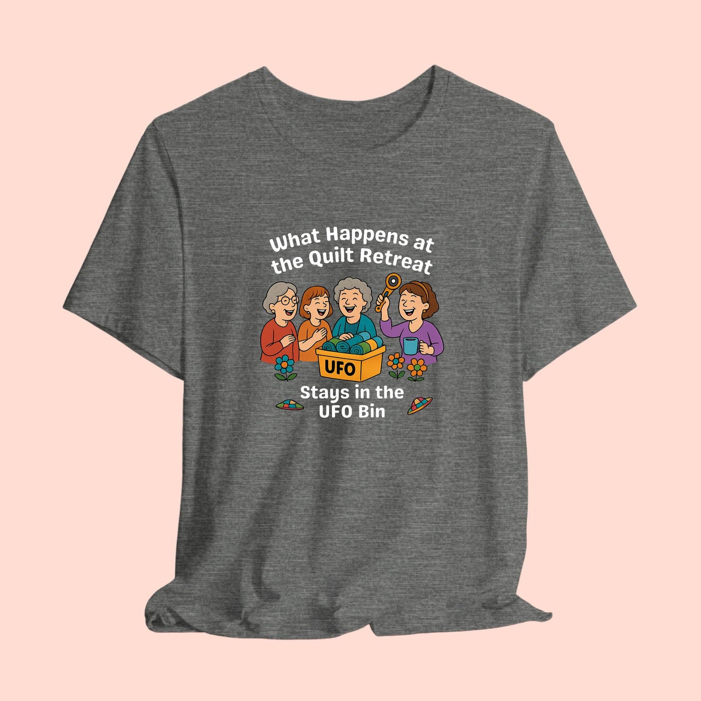A A t-shirt featuring a cartoon design of four laughing women at a quilt retreat with a bin labeled “UFO” full of fabric. Surrounded by patchwork flying saucers and flowers. Text reads “What happens at the quilt retreat stays in the UFO bin.”