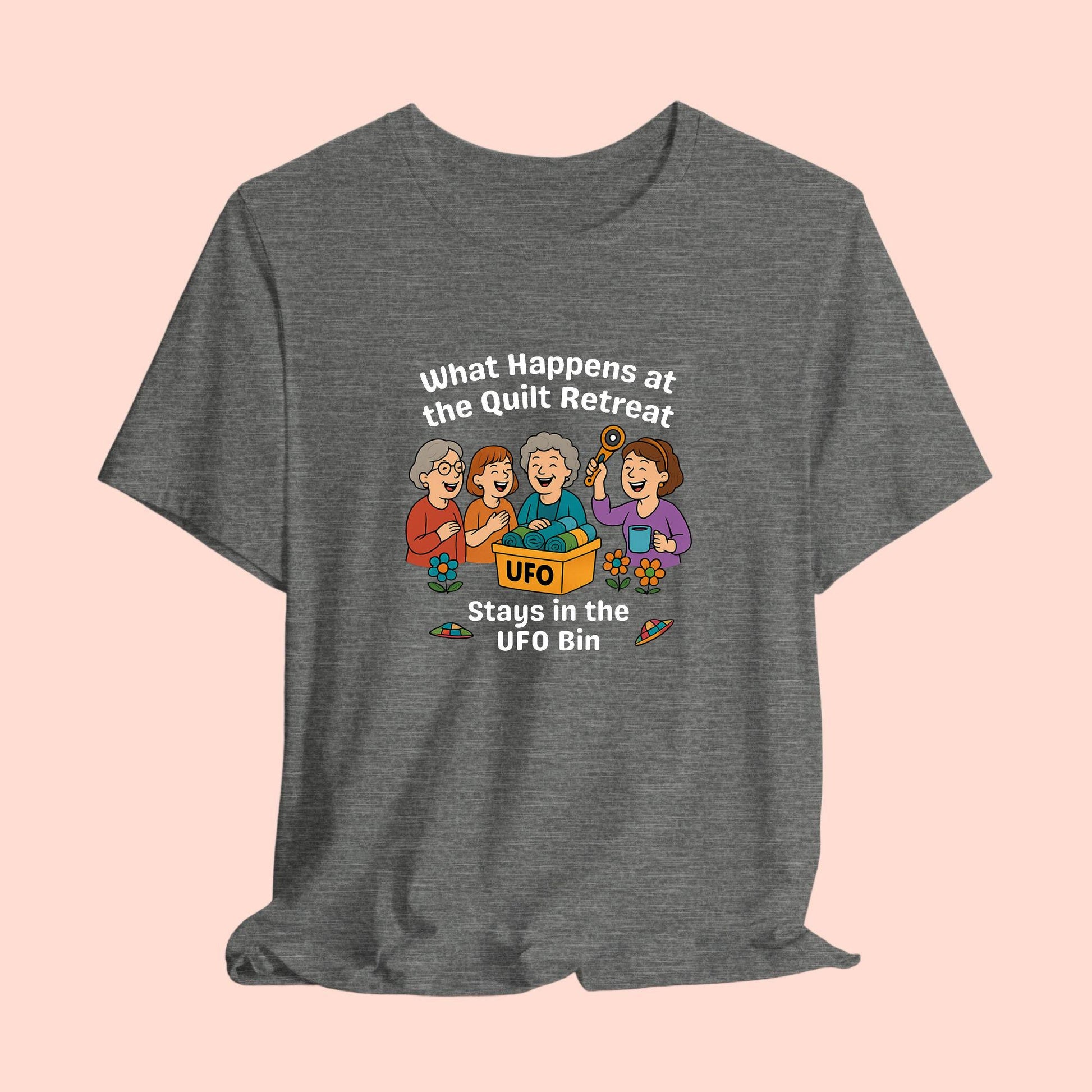 A A t-shirt featuring a cartoon design of four laughing women at a quilt retreat with a bin labeled “UFO” full of fabric. Surrounded by patchwork flying saucers and flowers. Text reads “What happens at the quilt retreat stays in the UFO bin.”