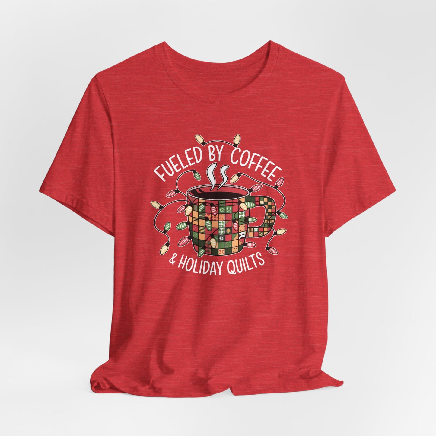 A Heather Red christmas quilting t-shirt featuring a cozy coffee mug covered in patchwork quilt patterns, wrapped in festive holiday lights, with the phrase "Fueled by Coffee & Holiday Quilts" surrounding the design.