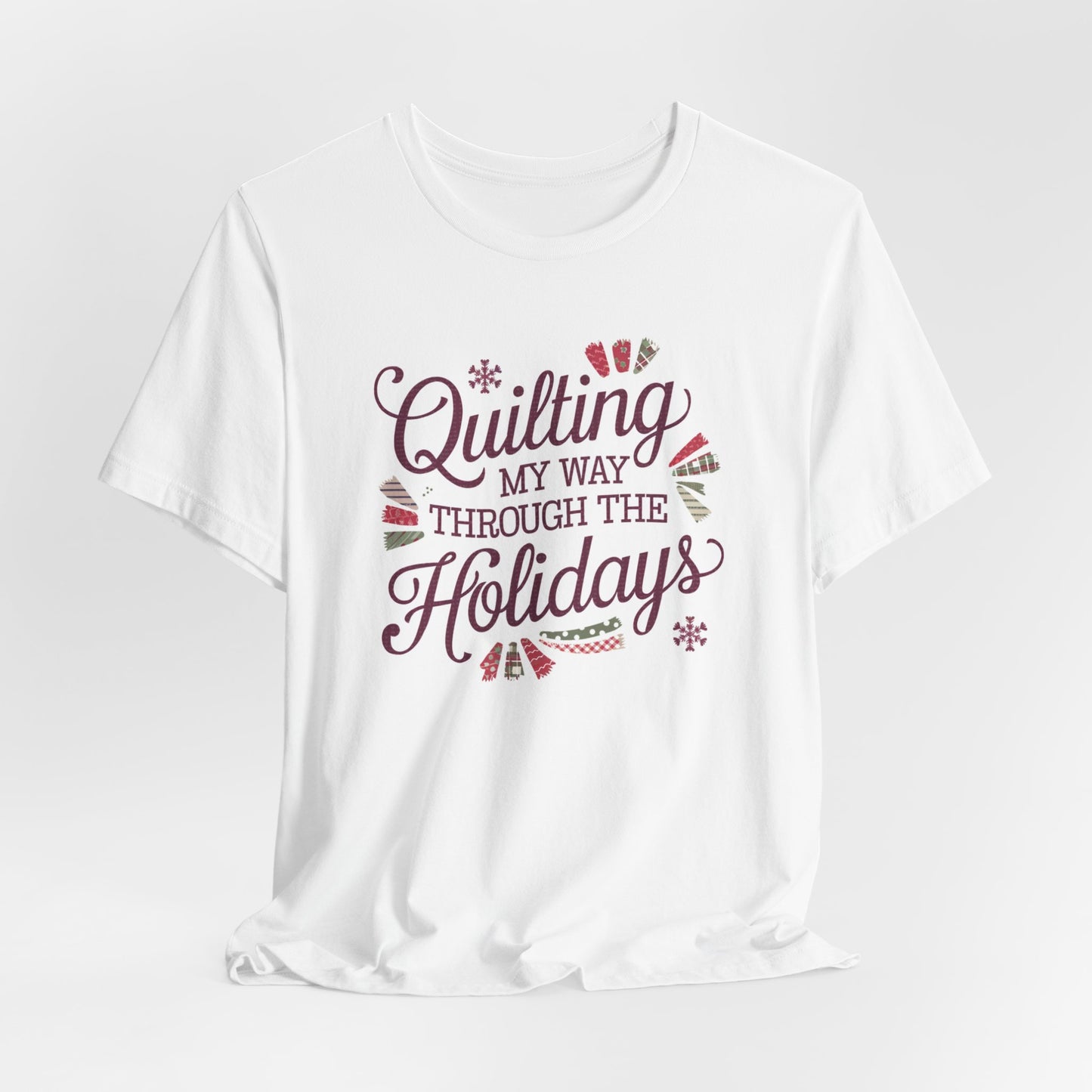 A White playful holiday-themed t-shirt with the phrase "Quilting My Way Through the Holidays" surrounded by quilting-inspired decorations like fabric swatches and snowflakes.