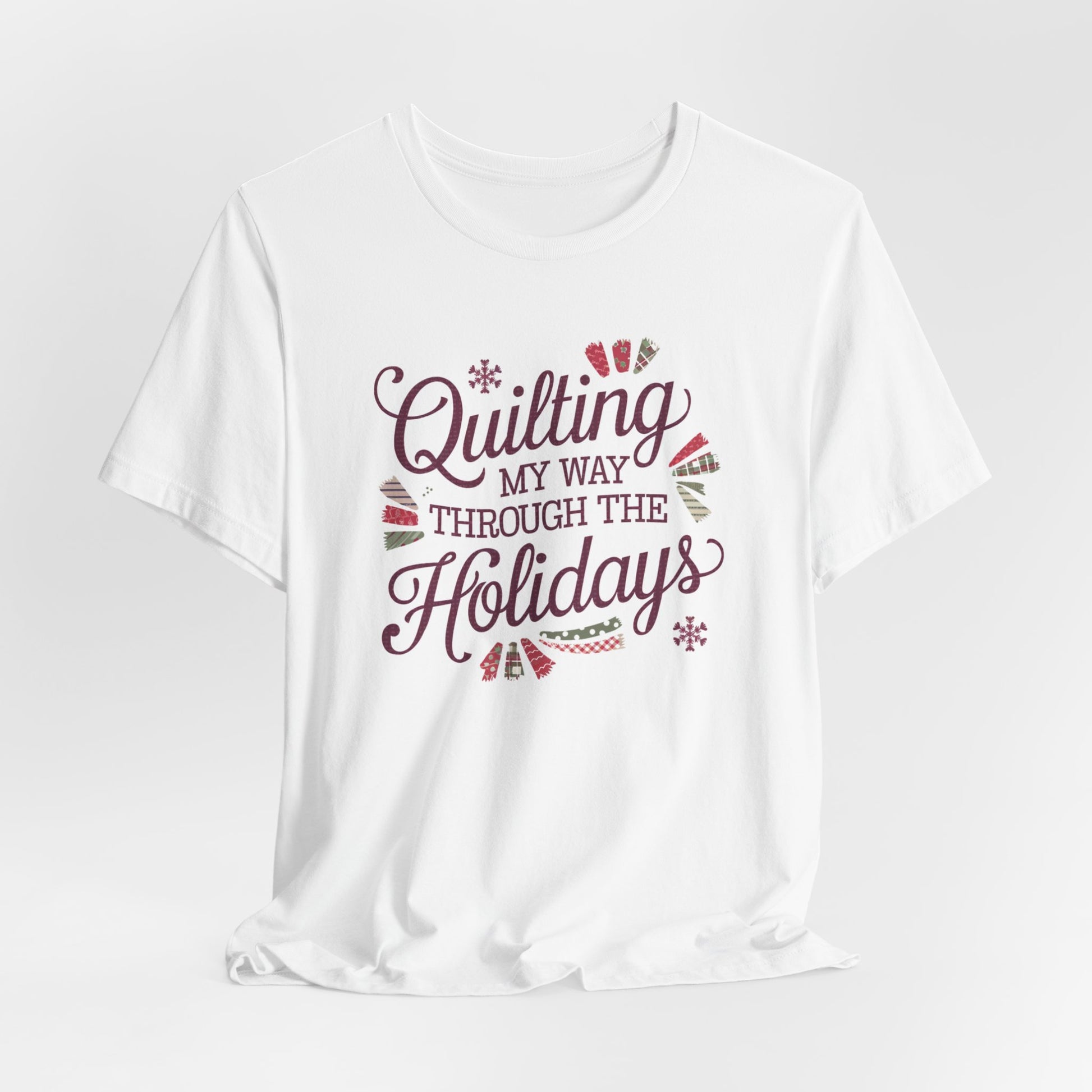 A White playful holiday-themed t-shirt with the phrase "Quilting My Way Through the Holidays" surrounded by quilting-inspired decorations like fabric swatches and snowflakes.