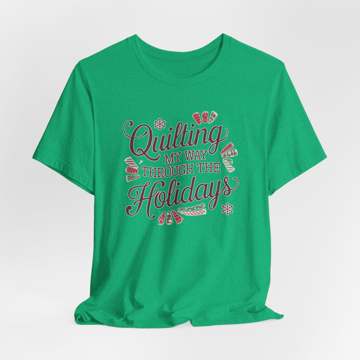 A Heather Kelly playful holiday-themed t-shirt with the phrase "Quilting My Way Through the Holidays" surrounded by quilting-inspired decorations like fabric swatches and snowflakes.