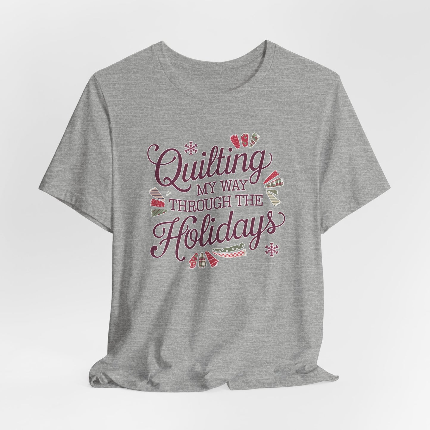 An Athletic Heather playful holiday-themed t-shirt with the phrase "Quilting My Way Through the Holidays" surrounded by quilting-inspired decorations like fabric swatches and snowflakes.