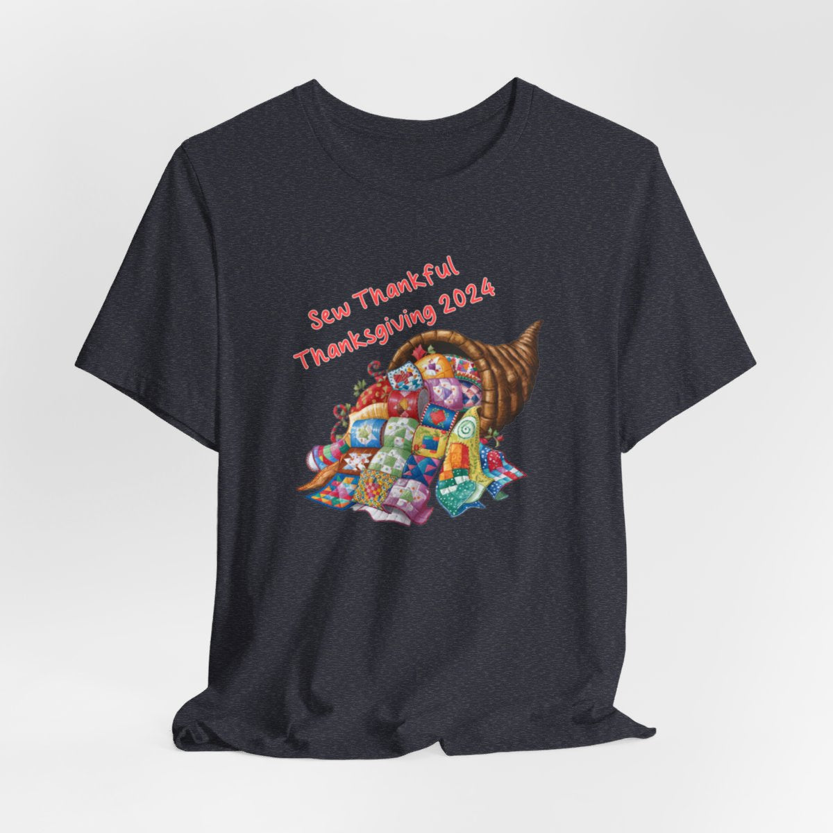A Heather Navy thanksgiving quilting t-shirt featuring a colorful quilted cornucopia overflowing with various quilt patterns and designs. Text reads "Sew Thankful Thanksgiving 2024" in red. Vibrant illustration combines quilting motifs with Thanksgiving imagery.