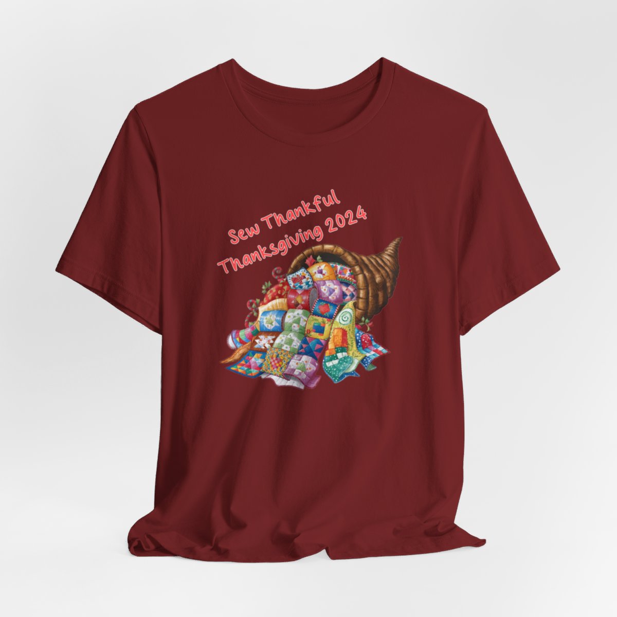 A Cardinal thanksgiving quilting t-shirt featuring a colorful quilted cornucopia overflowing with various quilt patterns and designs. Text reads "Sew Thankful Thanksgiving 2024" in red. Vibrant illustration combines quilting motifs with Thanksgiving imagery.