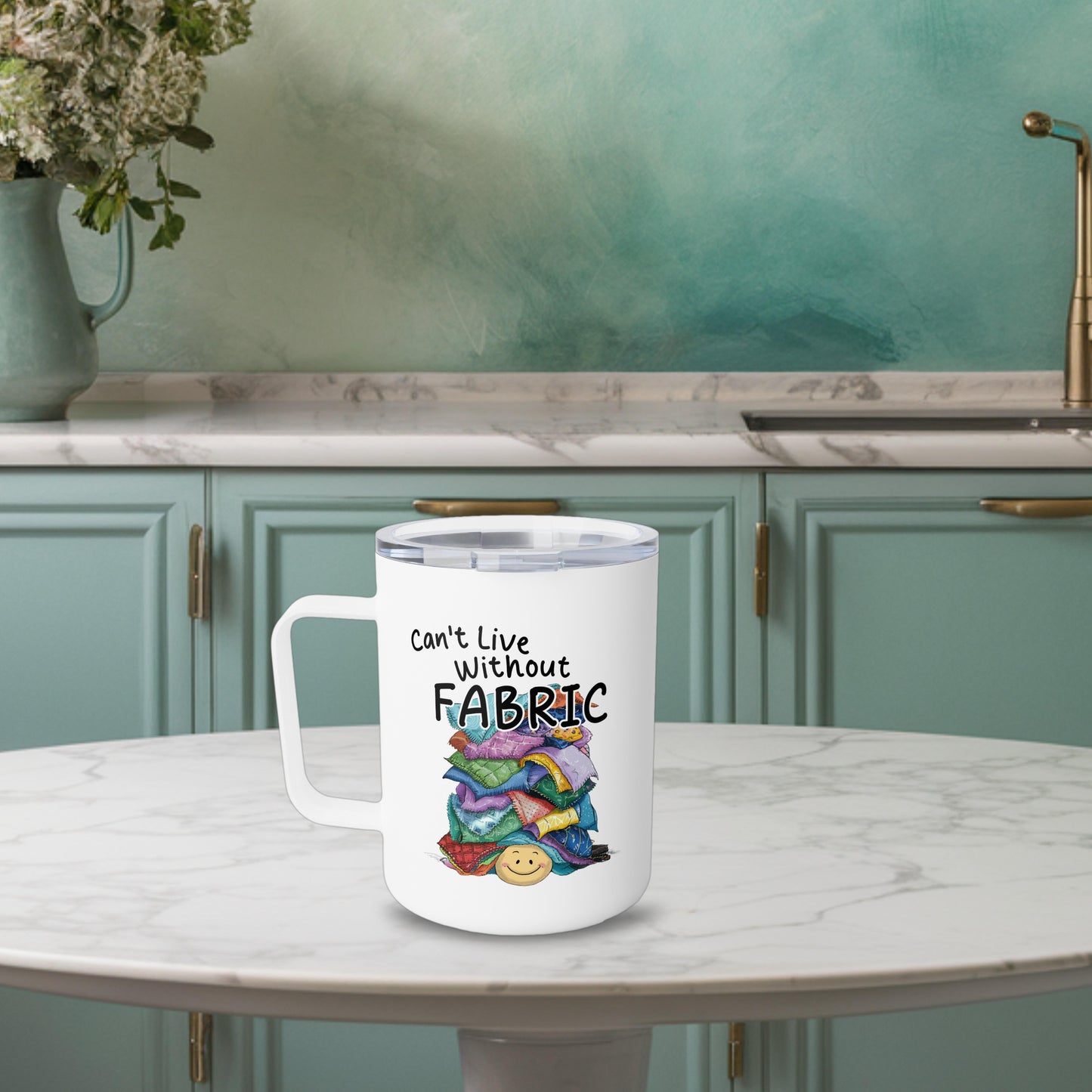 A funny quilting-inspired Insulated Mug with an Illustration of a smiling face underneath a pile of colorful fabric scraps. Text reads "Can't Live Without Fabric" in a handwritten style. The fabrics feature various patterns and textures in multiple colors.