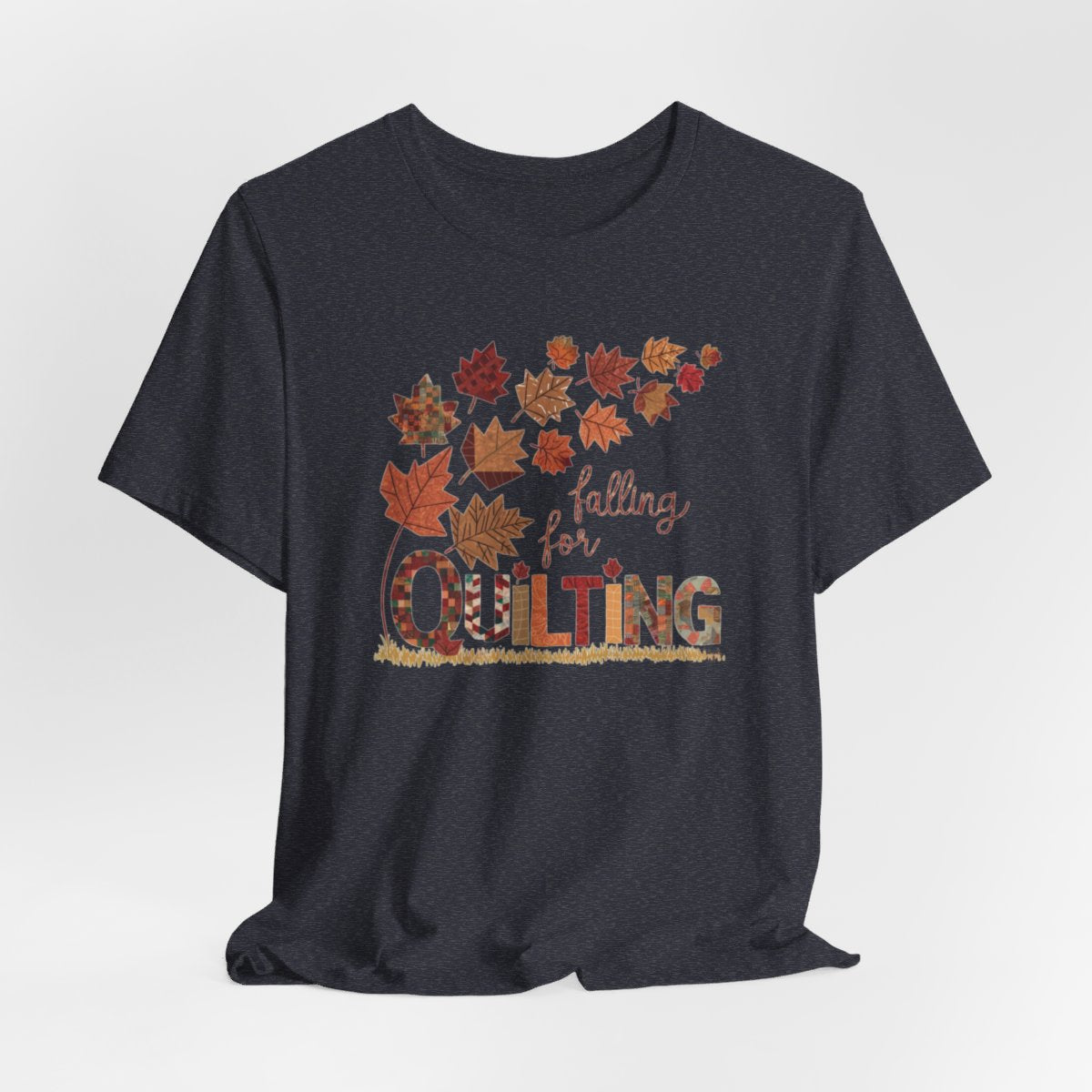 A Heather Navy thanksgiving quilting t-shirt featuring autumn leaves with various quilting patterns inside. Text reads "falling for QUILTING" with letters in patchwork style. Warm fall colors and quilting motifs combine in a seasonal design.