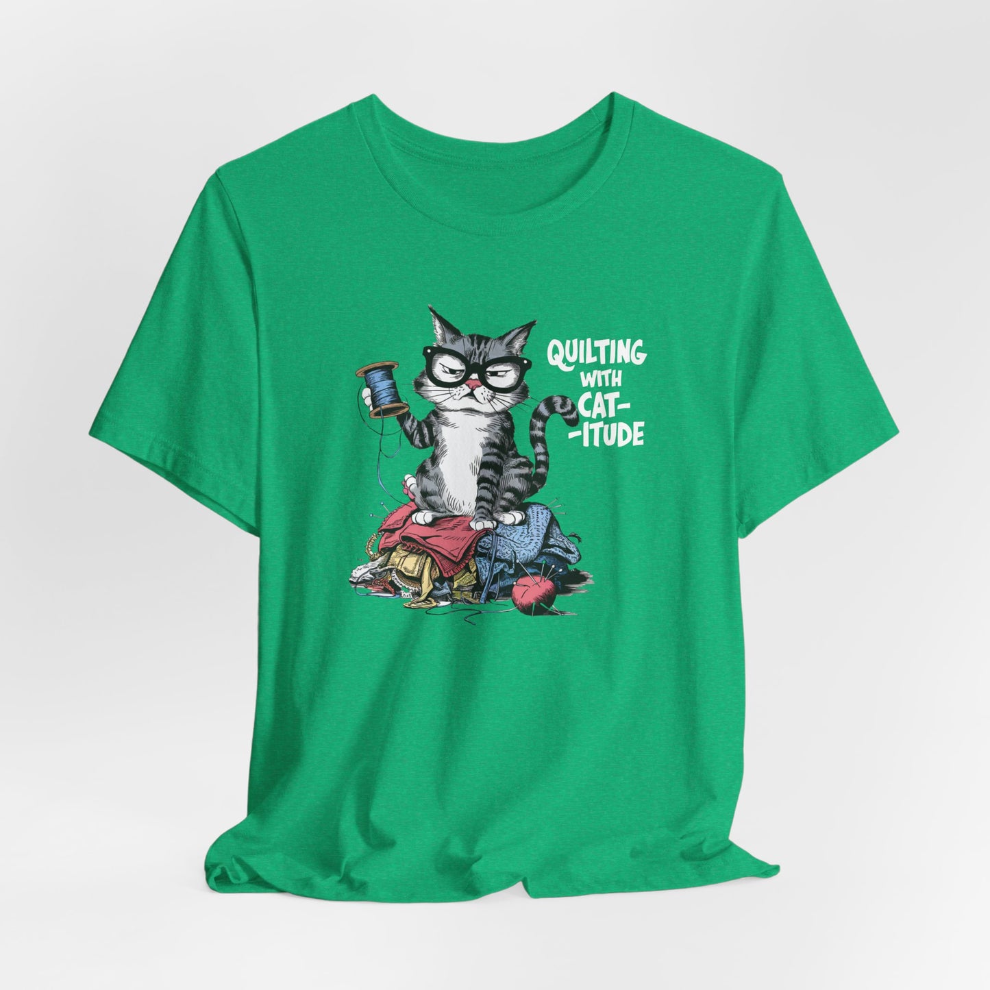 A Heather Kelly t-shirt featuring a sassy cartoon cat sitting on a pile of colorful fabric, holding a spool of thread, with the phrase 'Quilting with Cat-itude,' perfect for quilters and cat lovers.