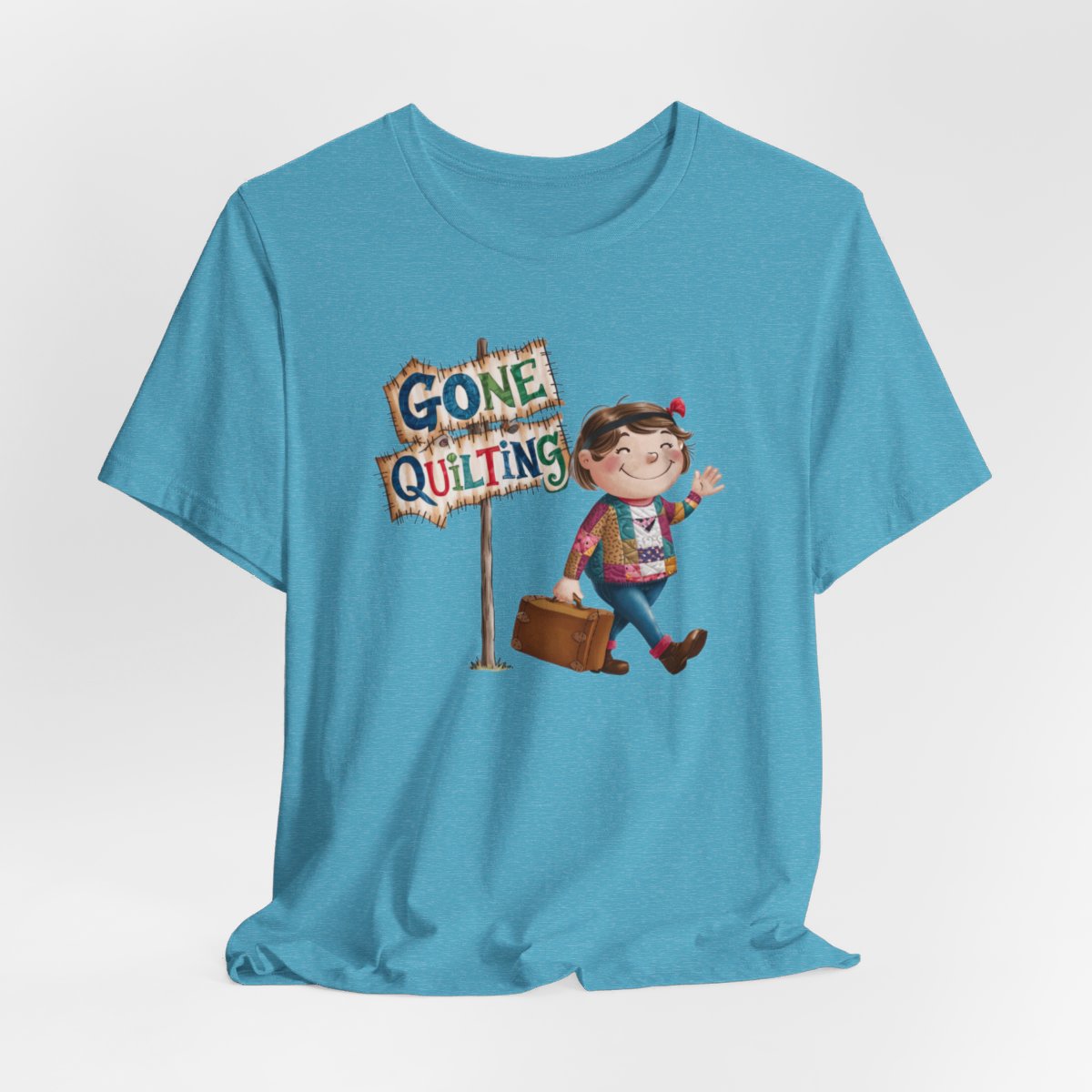 A Heather Aquafun quilting T-Shirt with a cartoon illustration of a smiling girl wearing a patchwork jacket, carrying a suitcase next to a wooden sign that reads "Gone Quilting" in colorful letters.