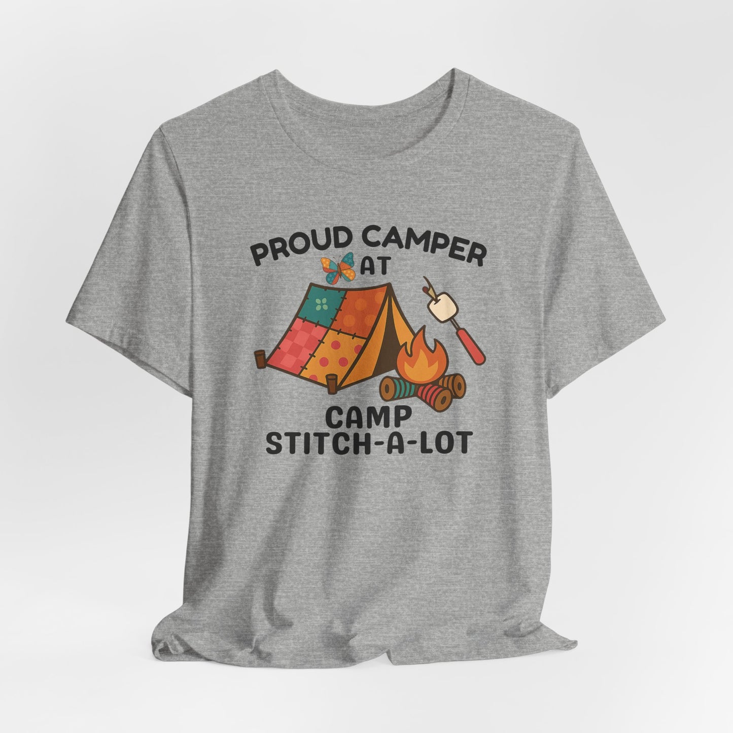 A An Athletic Heather t-shirt featuring a colorful design with a patchwork tent, thread spool campfire, marshmallow on a seam ripper stick, and a butterfly. Text reads "Proud Camper at Camp Stitch-a-Lot." Fun gift for quilting retreats.