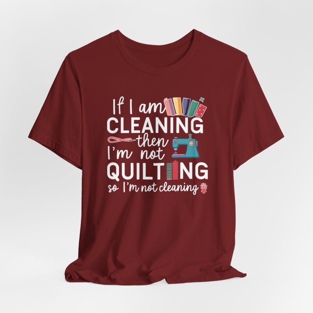 A Cardinal funny quilting T-Shirt with the phrase If I am Cleaning then I'm not Quilting in a bold and cheerful font with images of quilting icons and supplies