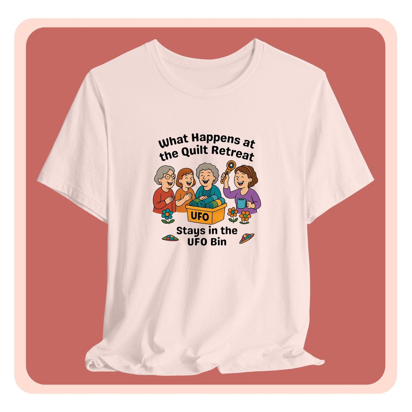 A A t-shirt featuring a cartoon design of four laughing women at a quilt retreat with a bin labeled “UFO” full of fabric. Surrounded by patchwork flying saucers and flowers. Text reads “What happens at the quilt retreat stays in the UFO bin.”