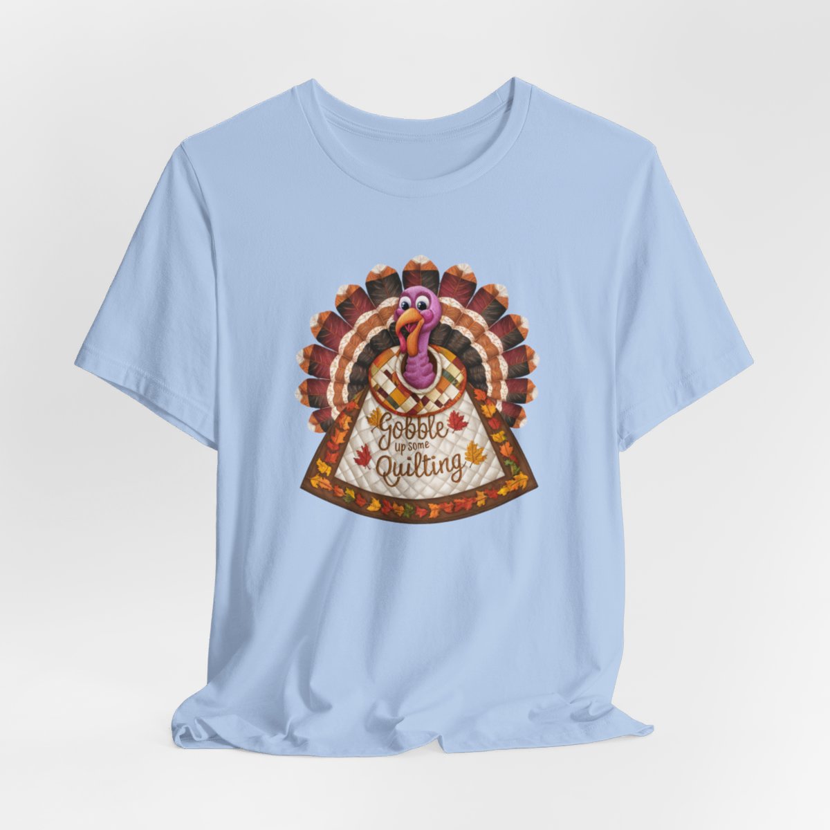A Baby Blue thanksgiving quilting t-shirt featuring a quilted turkey design with colorful patchwork feathers and body. Text on turkey reads "Gobble up some Quilting" surrounded by fall leaves. Combines Thanksgiving imagery with quilting motifs in a whimsical illustration.