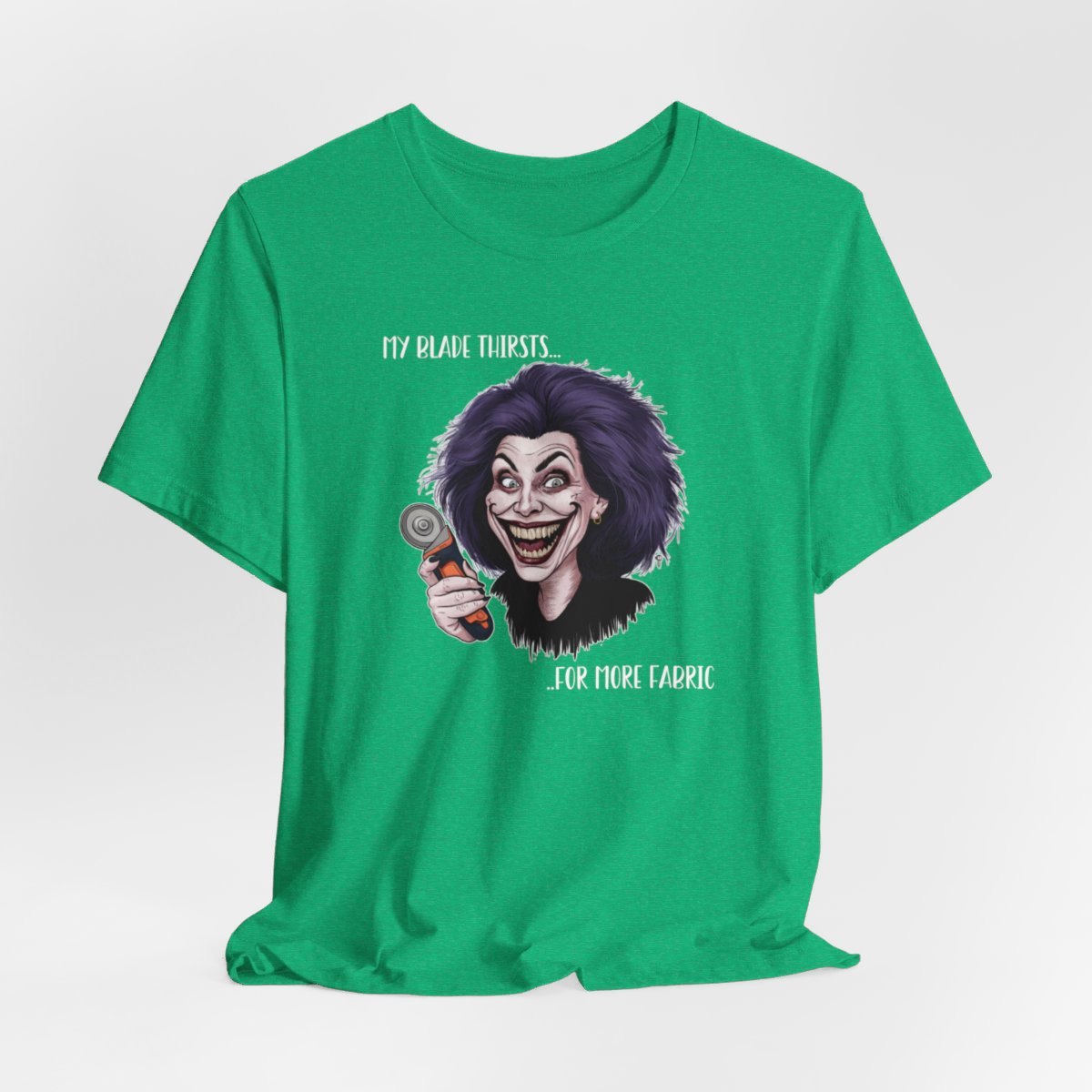 A Heather Kelly T-shirt showing a cartoonish character with wild purple hair and an exaggerated grin, holding a rotary cutter. Text reads "My blade thirsts... for more fabric", humorously depicting a quilter's endless desire for new materials.