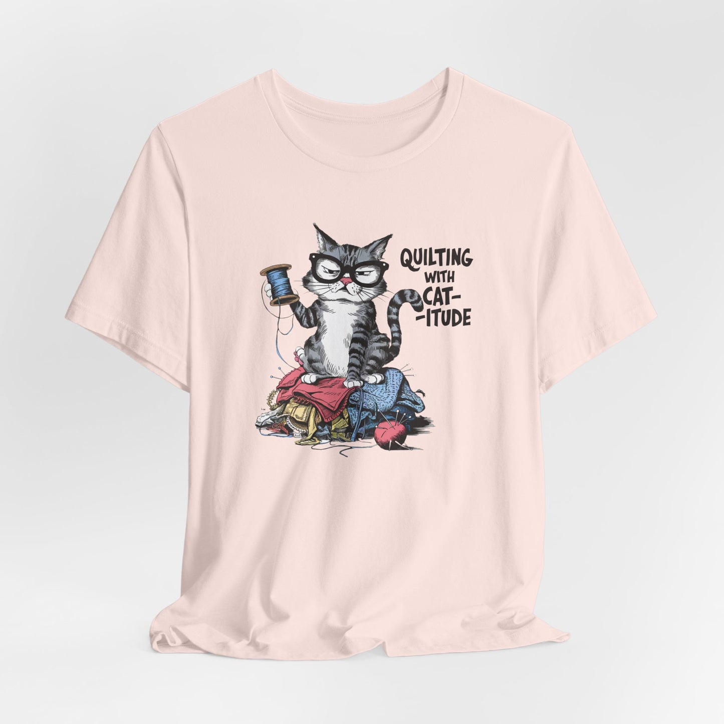 A Soft Pink t-shirt featuring a sassy cartoon cat sitting on a pile of colorful fabric, holding a spool of thread, with the phrase 'Quilting with Cat-itude,' perfect for quilters and cat lovers.