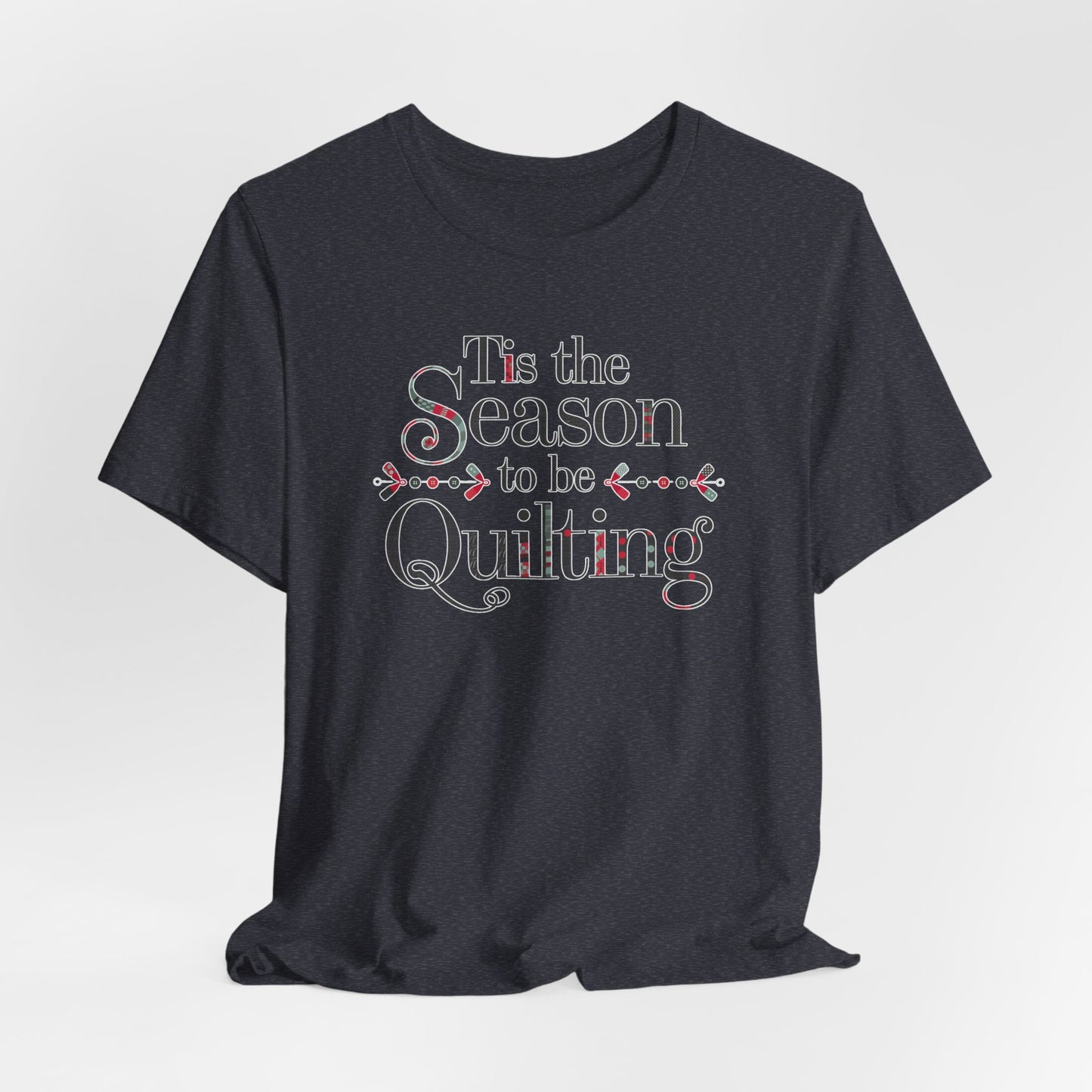 A Heather Navy festive t-shirt featuring the phrase "'Tis the Season to be Quilting," with playful sewing-inspired elements like needles, buttons, and fabric patterns integrated into the typography.
