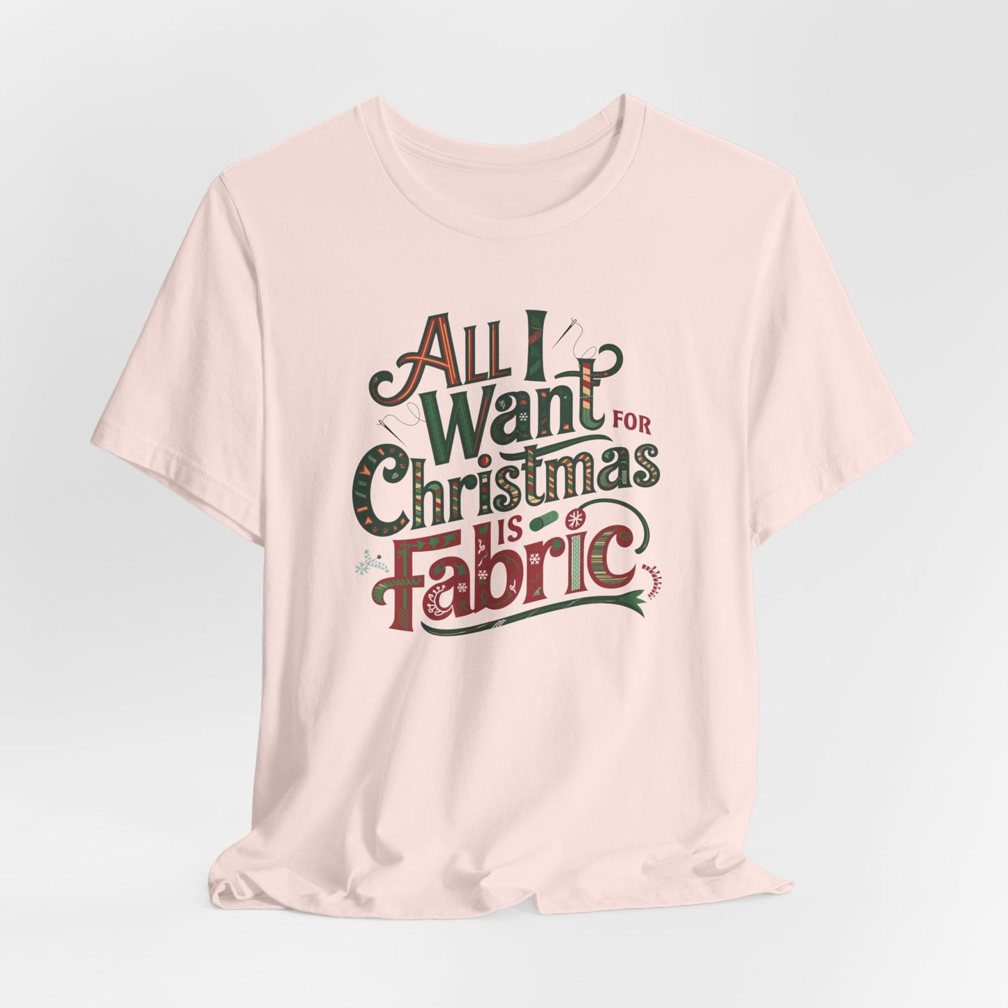 A Soft Pink festive quilting christmas T-shirt featuring the phrase "All I Want for Christmas is Fabric," with fun sewing and quilting accents like needles and threads swirling around the text.