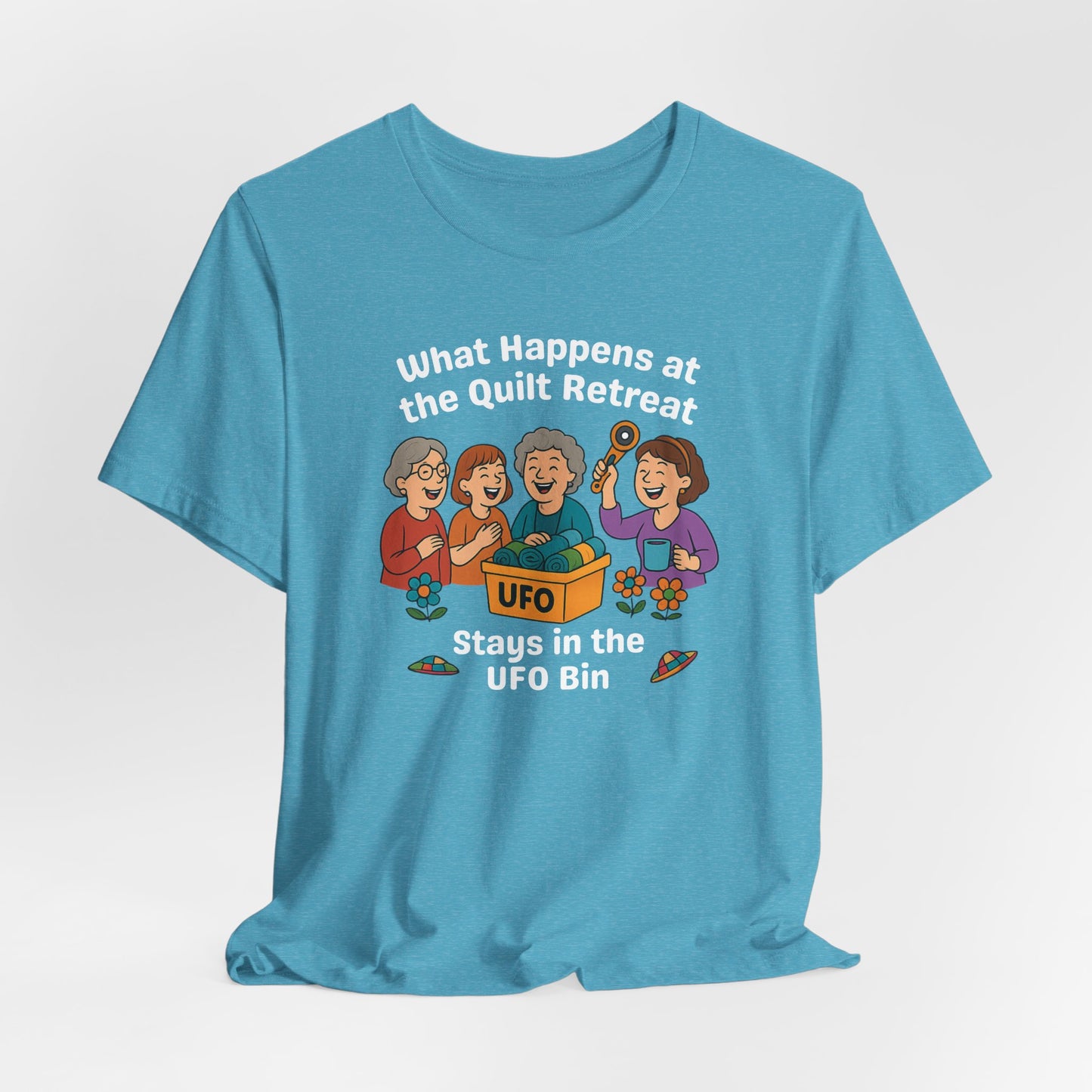 A A Heather Aqua t-shirt featuring a cartoon design of four laughing women at a quilt retreat with a bin labeled “UFO” full of fabric. Surrounded by patchwork flying saucers and flowers. Text reads “What happens at the quilt retreat stays in the UFO bin.”