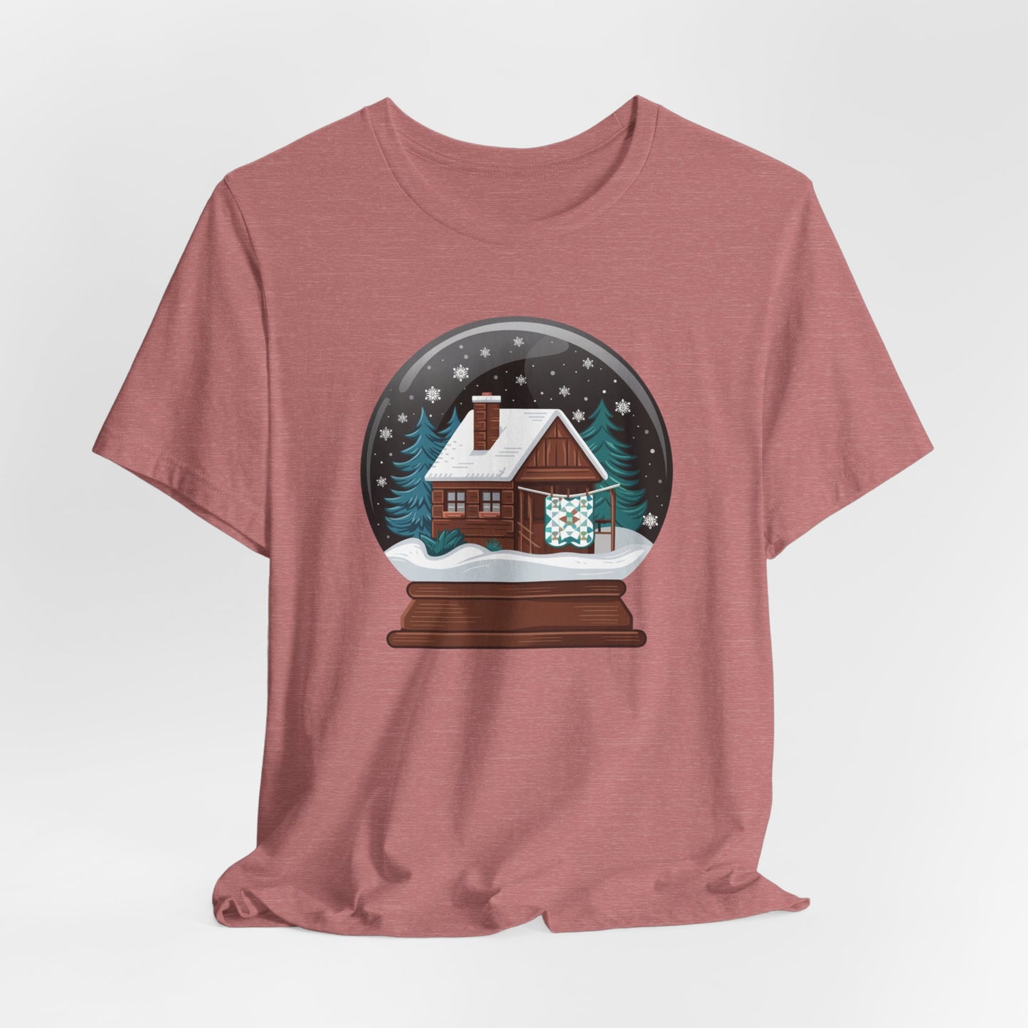 A Heather Mauve christmas quilting t-shirt featuring a cozy log cabin covered in snow, with a quilt hanging outside, set inside a snow globe with swirling snowflakes, blending quilting and holiday warmth.