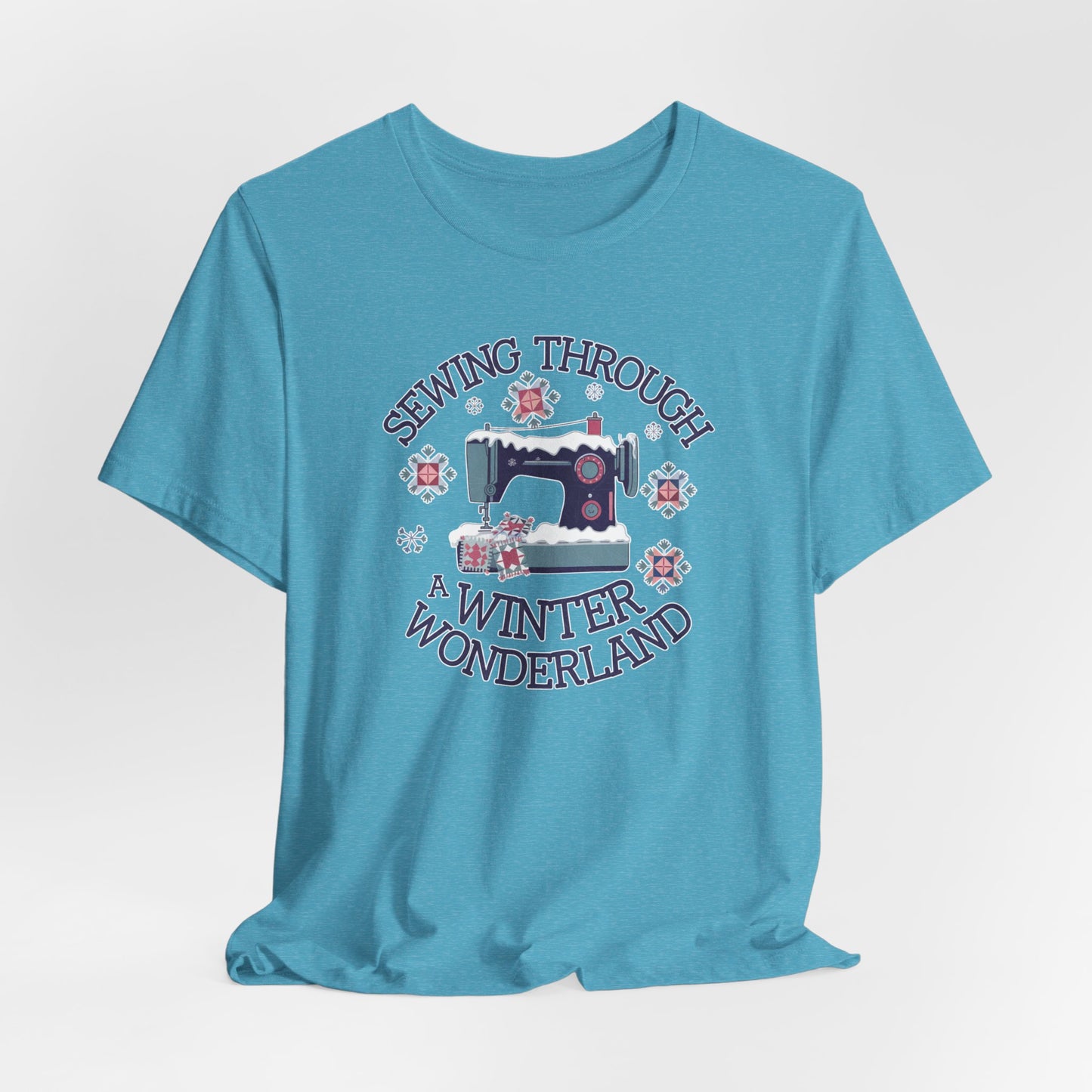 A Heather Aqua winter quilting t-shirt featuring a sewing machine covered in snow, surrounded by quilted snowflakes, with the phrase "Sewing Through a Winter Wonderland" above and below the image.
