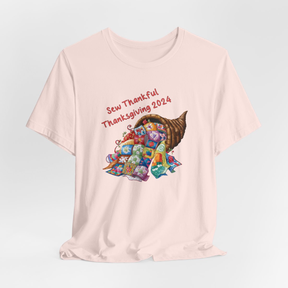 A Soft Pink thanksgiving quilting t-shirt featuring a colorful quilted cornucopia overflowing with various quilt patterns and designs. Text reads "Sew Thankful Thanksgiving 2024" in red. Vibrant illustration combines quilting motifs with Thanksgiving imagery.