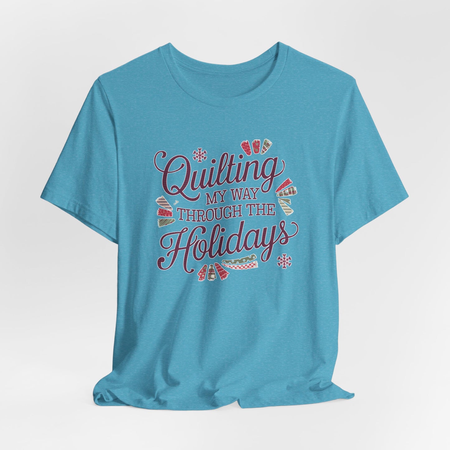 A Heather Aqua playful holiday-themed t-shirt with the phrase "Quilting My Way Through the Holidays" surrounded by quilting-inspired decorations like fabric swatches and snowflakes.