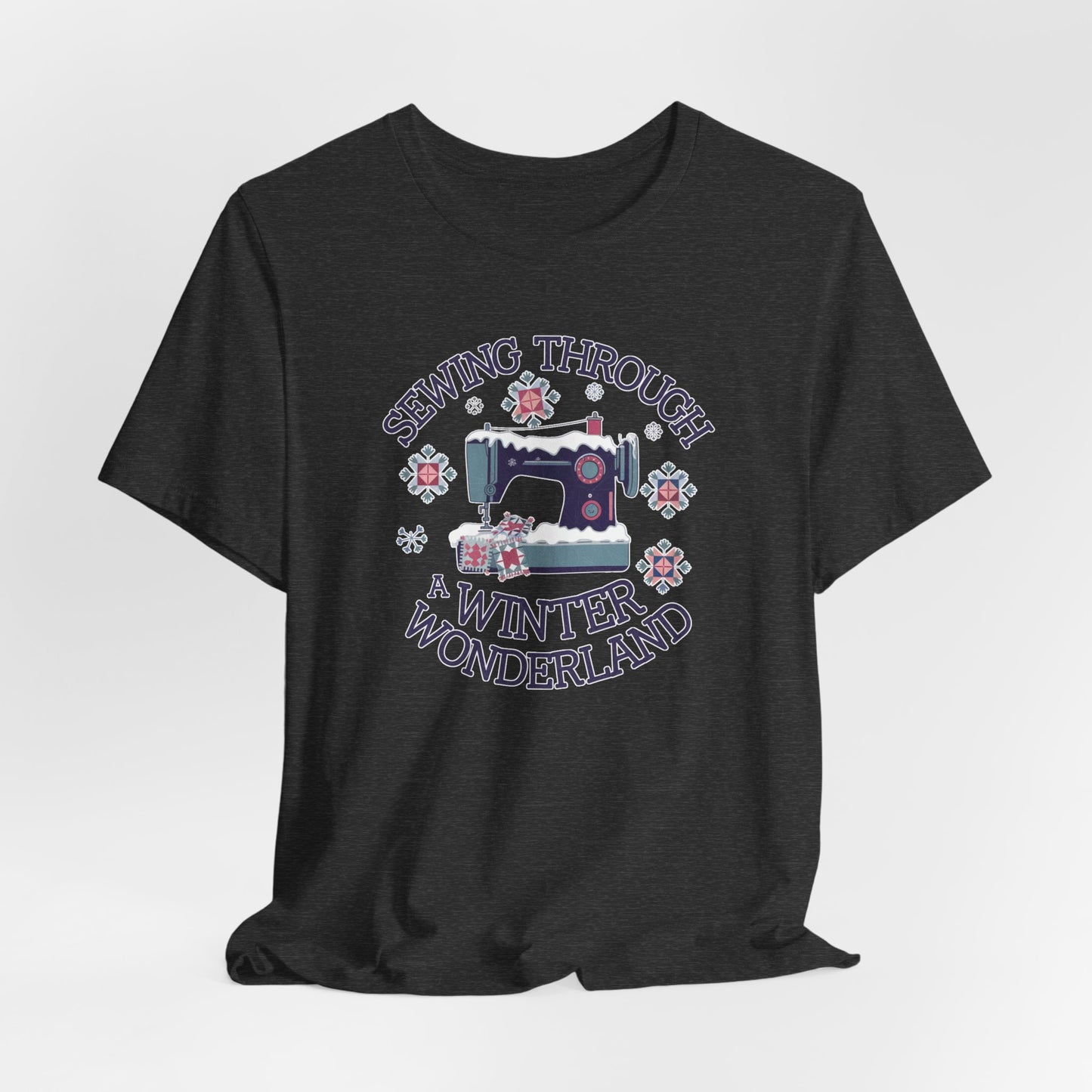 A Dark Grey Heather winter quilting t-shirt featuring a sewing machine covered in snow, surrounded by quilted snowflakes, with the phrase "Sewing Through a Winter Wonderland" above and below the image.
