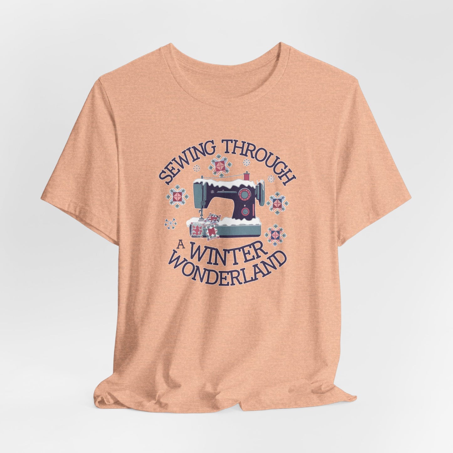 A Heather Peach winter quilting t-shirt featuring a sewing machine covered in snow, surrounded by quilted snowflakes, with the phrase "Sewing Through a Winter Wonderland" above and below the image.