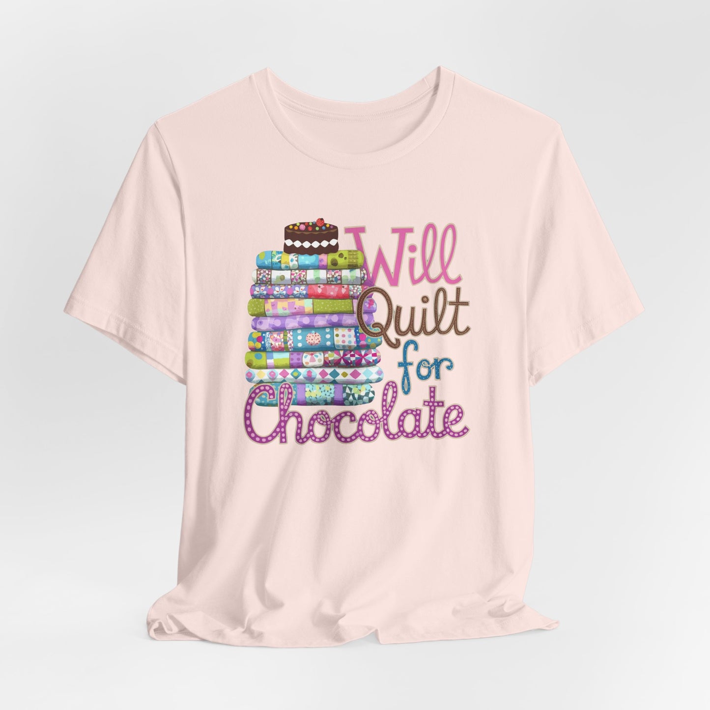 A Soft Pink t-shirt featuring a whimsical design of a colorful stack of quilts topped with a chocolate cake and the phrase 'Will Quilt for Chocolate,' perfect for quilters who love sweets.