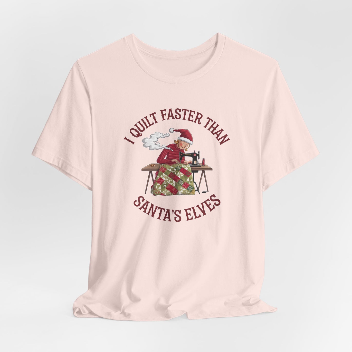 A Soft Pink christmas quilting t-shirt featuring a quilter working at a sewing machine, quilting a festive holiday quilt, with the phrase "I Quilt Faster Than Santa's Elves" above the image, highlighting the speed and skill of the quilter.