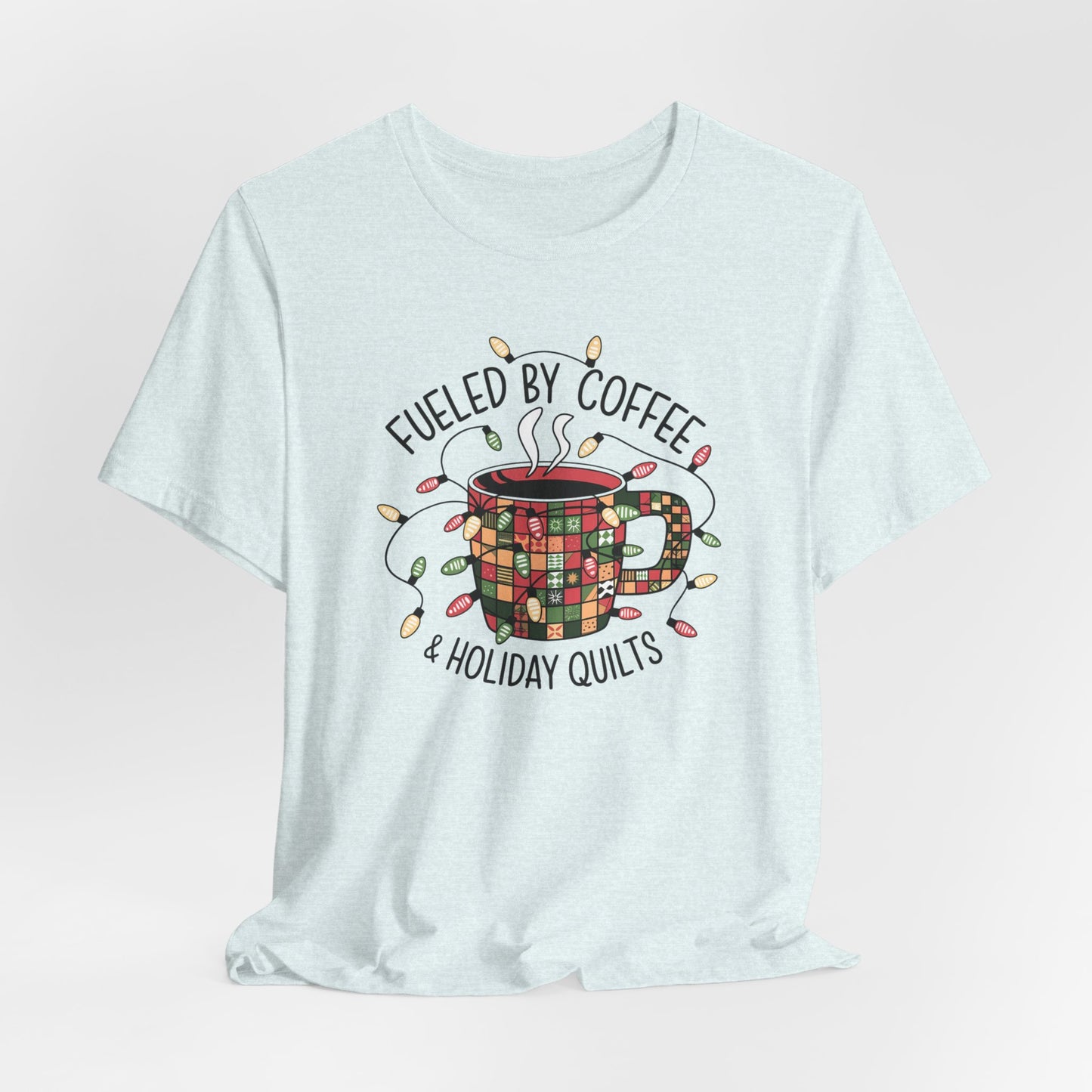 A Heather Ice Blue christmas quilting t-shirt featuring a cozy coffee mug covered in patchwork quilt patterns, wrapped in festive holiday lights, with the phrase "Fueled by Coffee & Holiday Quilts" surrounding the design.