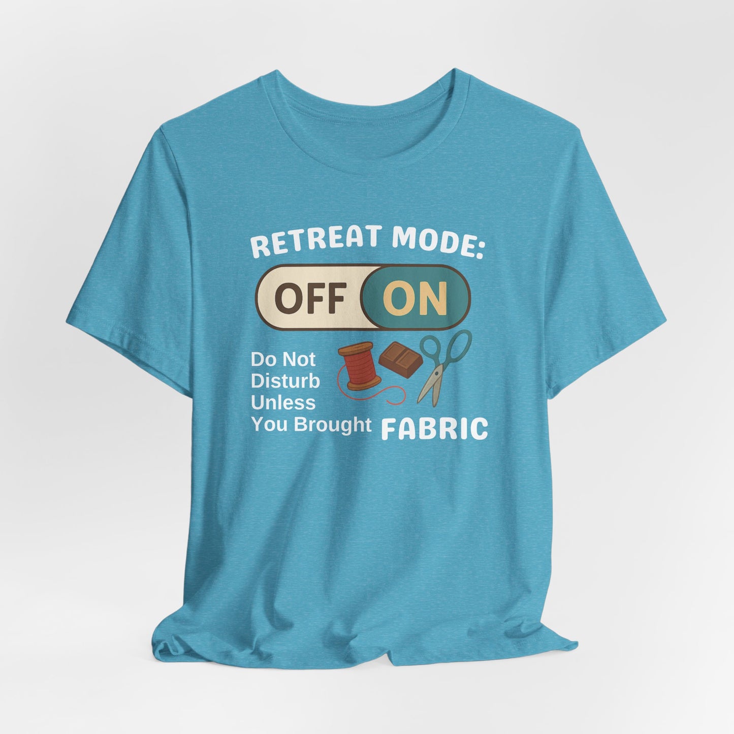 A A Heather Aqua t-shirt with a toggle switch that says "Retreat Mode: OFF/ON" surrounded by a spool of thread, chocolate, and scissors. Text below reads "Do not disturb unless you brought fabric." Fun gift for quilting retreats.
