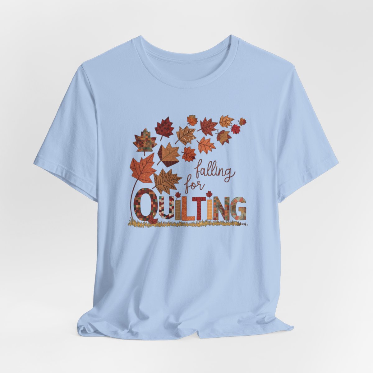 A Baby Blue thanksgiving quilting t-shirt featuring autumn leaves with various quilting patterns inside. Text reads "falling for QUILTING" with letters in patchwork style. Warm fall colors and quilting motifs combine in a seasonal design.
