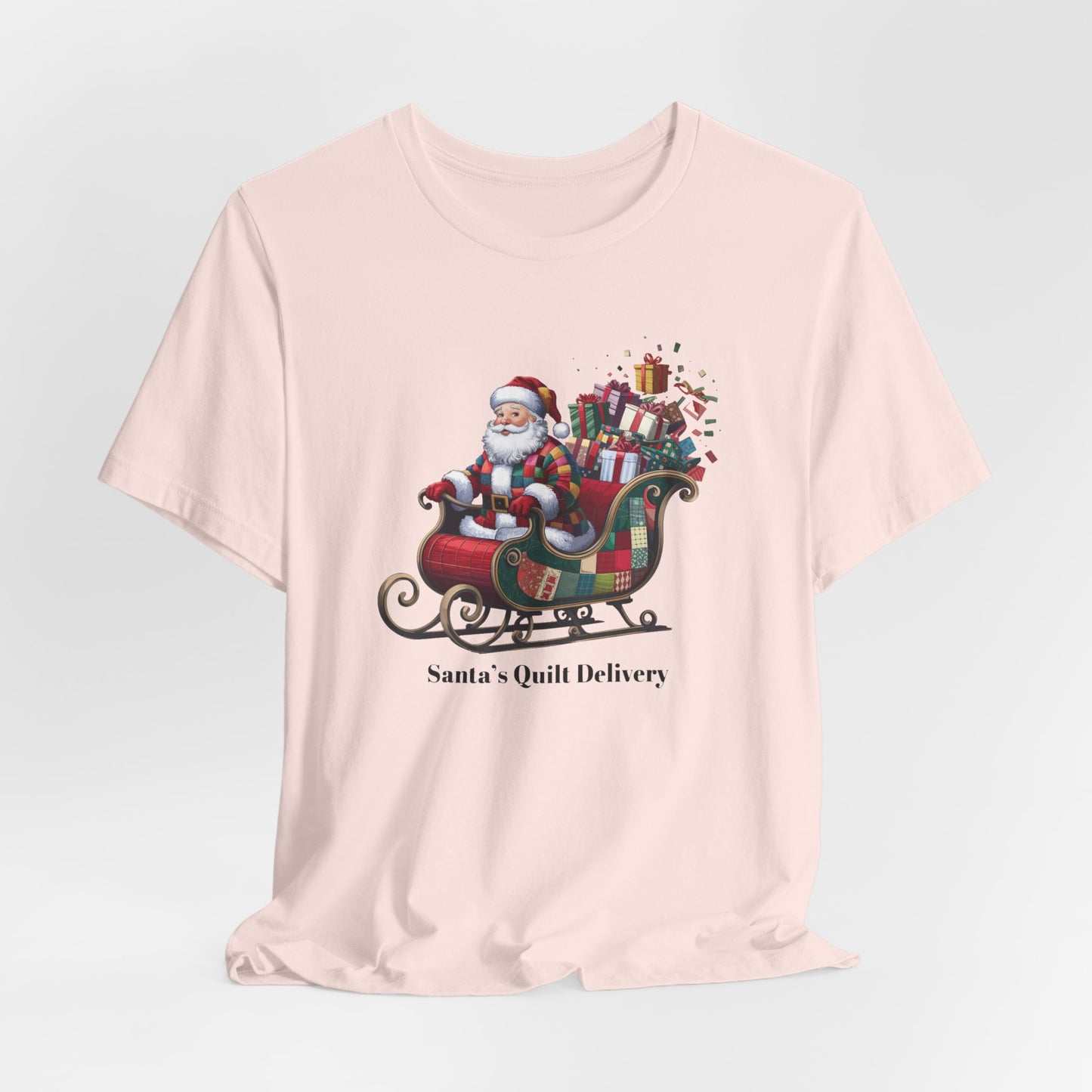 A Soft Pink christmas quilting t-shirt featuring Santa Claus wearing a patchwork quilt coat, sitting in a sleigh filled with gifts and quilts, with the phrase "Santa's Quilt Delivery" below, combining holiday cheer and quilting fun.