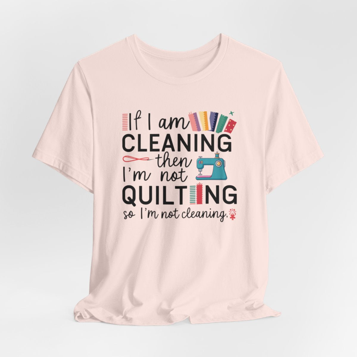 A Soft Pink funny quilting T-Shirt with the phrase If I am Cleaning then I'm not Quilting in a bold and cheerful font with images of quilting icons and supplies