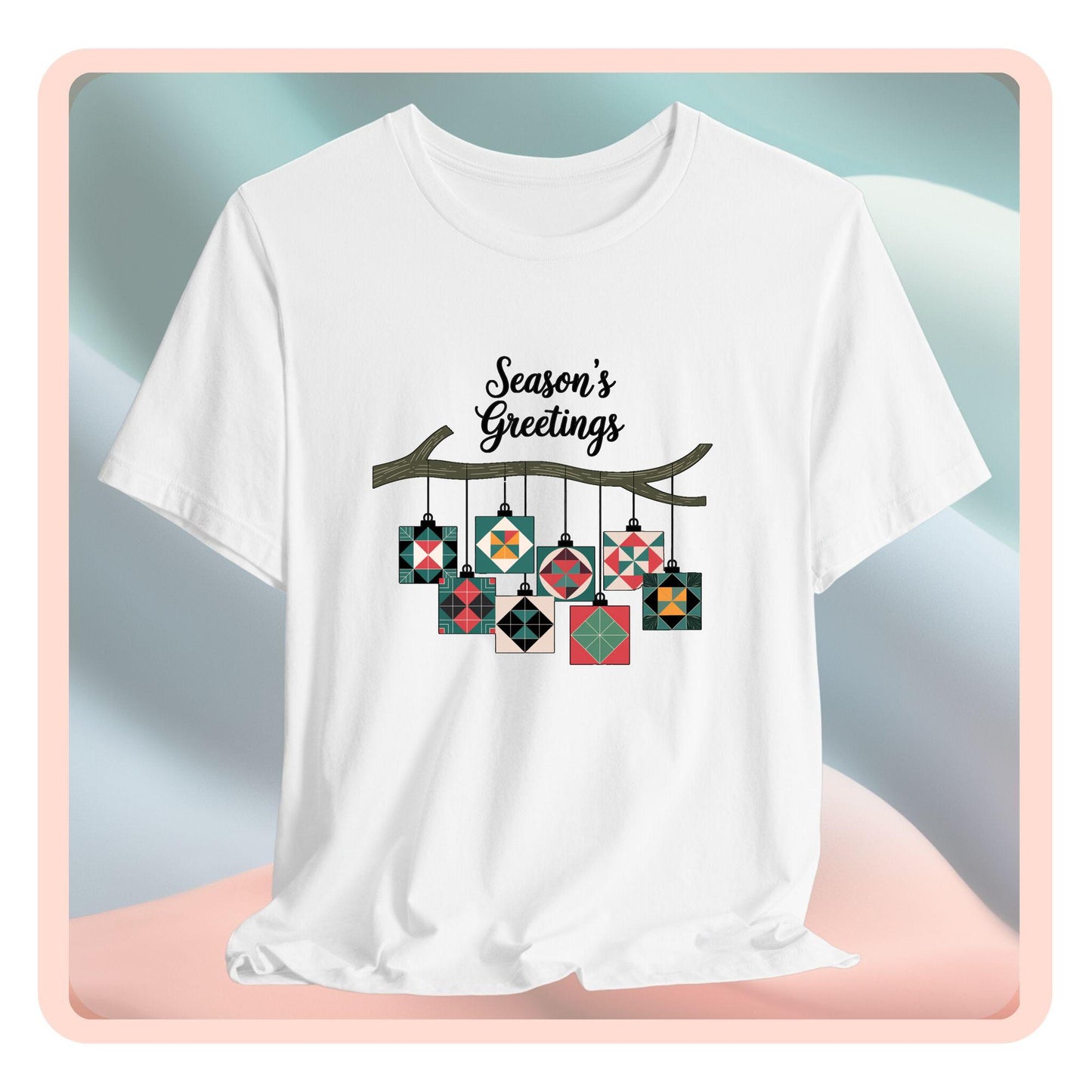 A christmas quilting t-shirt featuring quilt block ornaments hanging from a branch, featuring the phrase "Season's Greetings" above. The ornaments showcase various quilting patterns to highlight the holiday quilting theme.