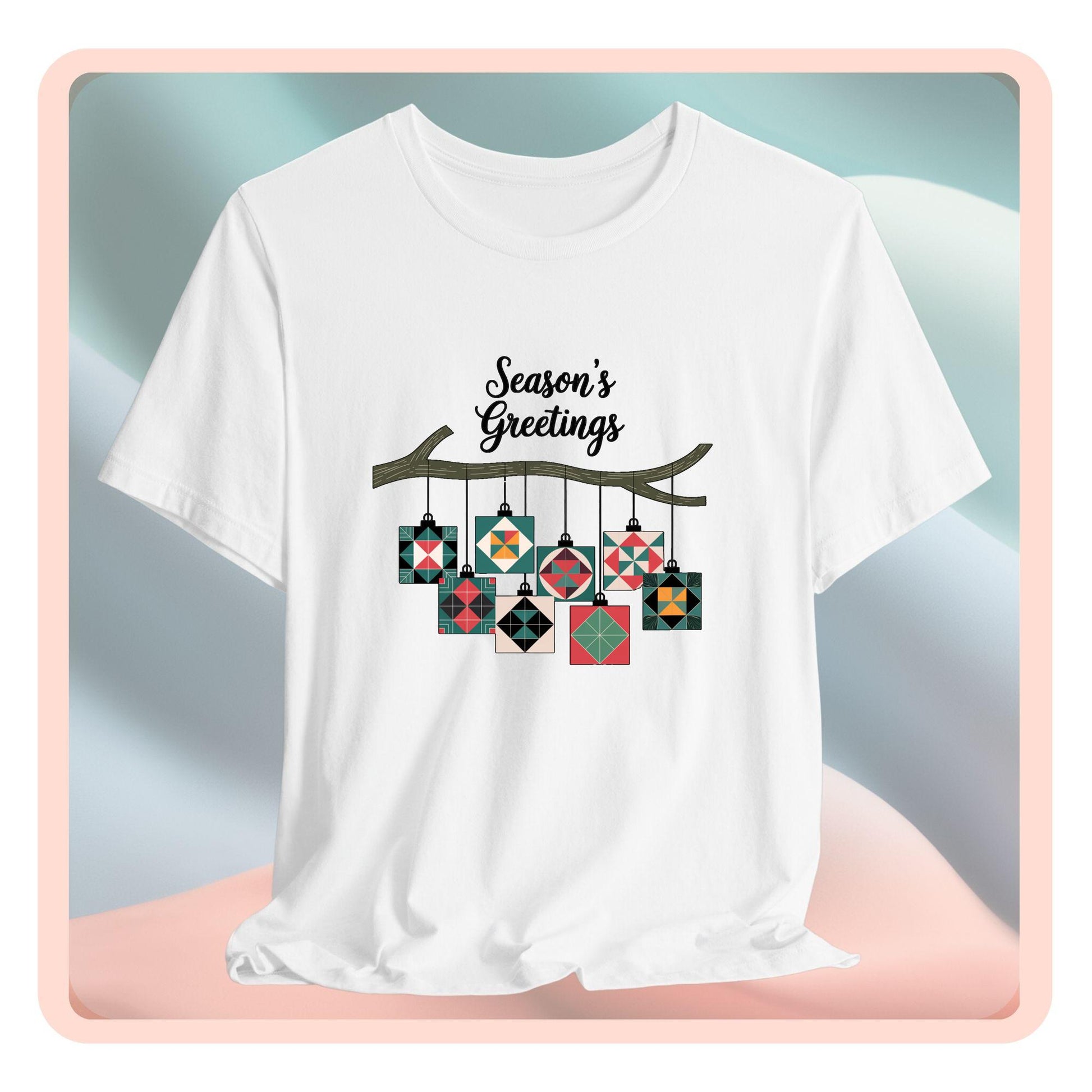 A christmas quilting t-shirt featuring quilt block ornaments hanging from a branch, featuring the phrase "Season's Greetings" above. The ornaments showcase various quilting patterns to highlight the holiday quilting theme.
