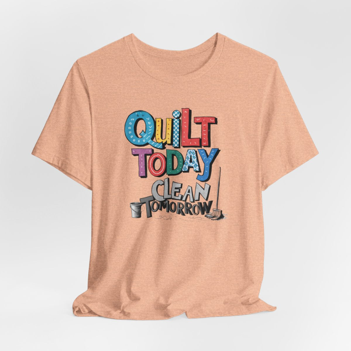 A Heather Peach funny quilting T-Shirt with the phrase Quilt Today, Clean Tomorrow in a bold and cheerful font with a broom off to the side